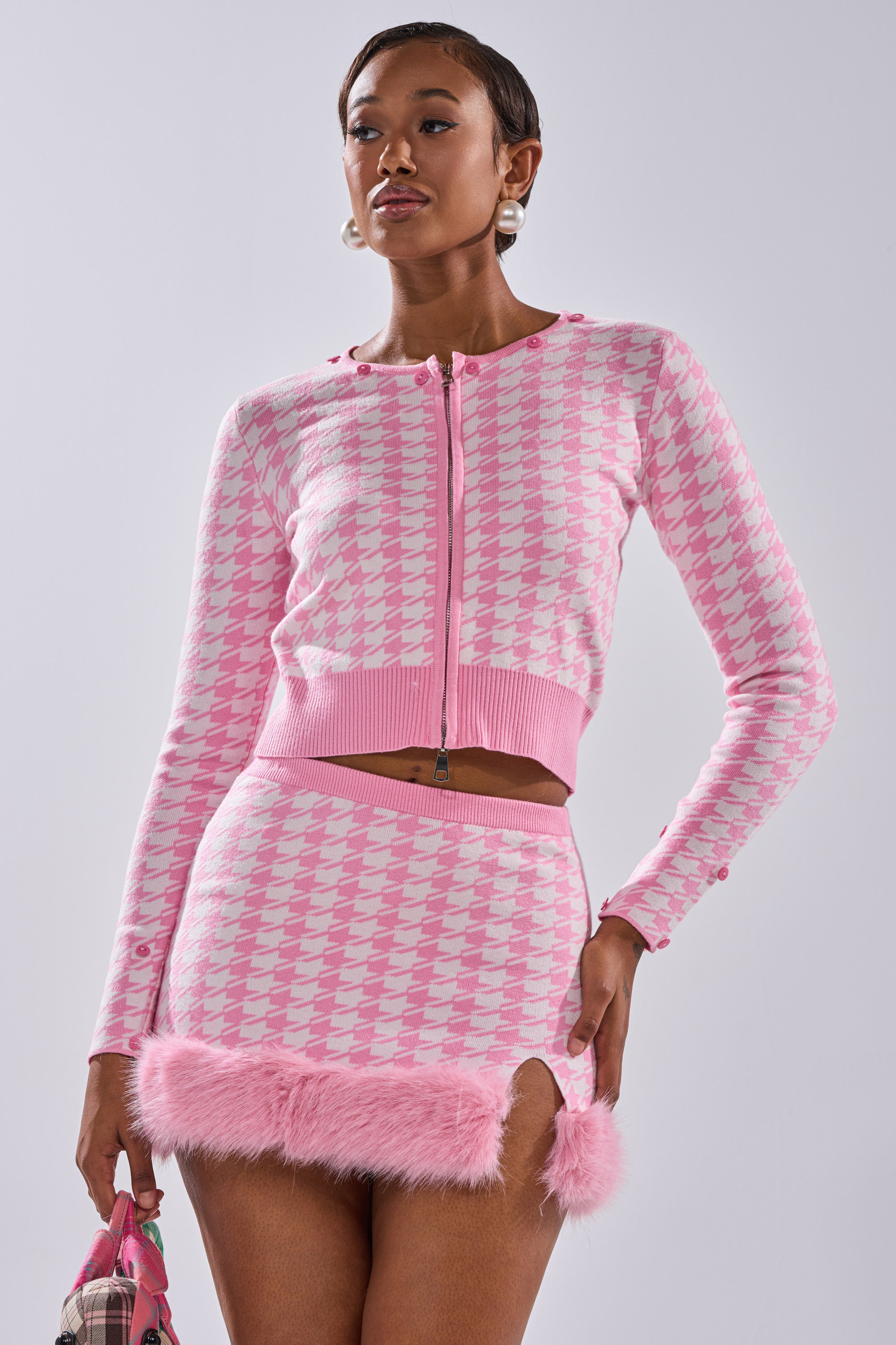 A woman poses confidently against a plain light background in the QUEEN OF LOVE SWEATER WITH FUR TRIM, styled with a houndstooth knit mini skirt and a small bag for a chic, coordinated look.