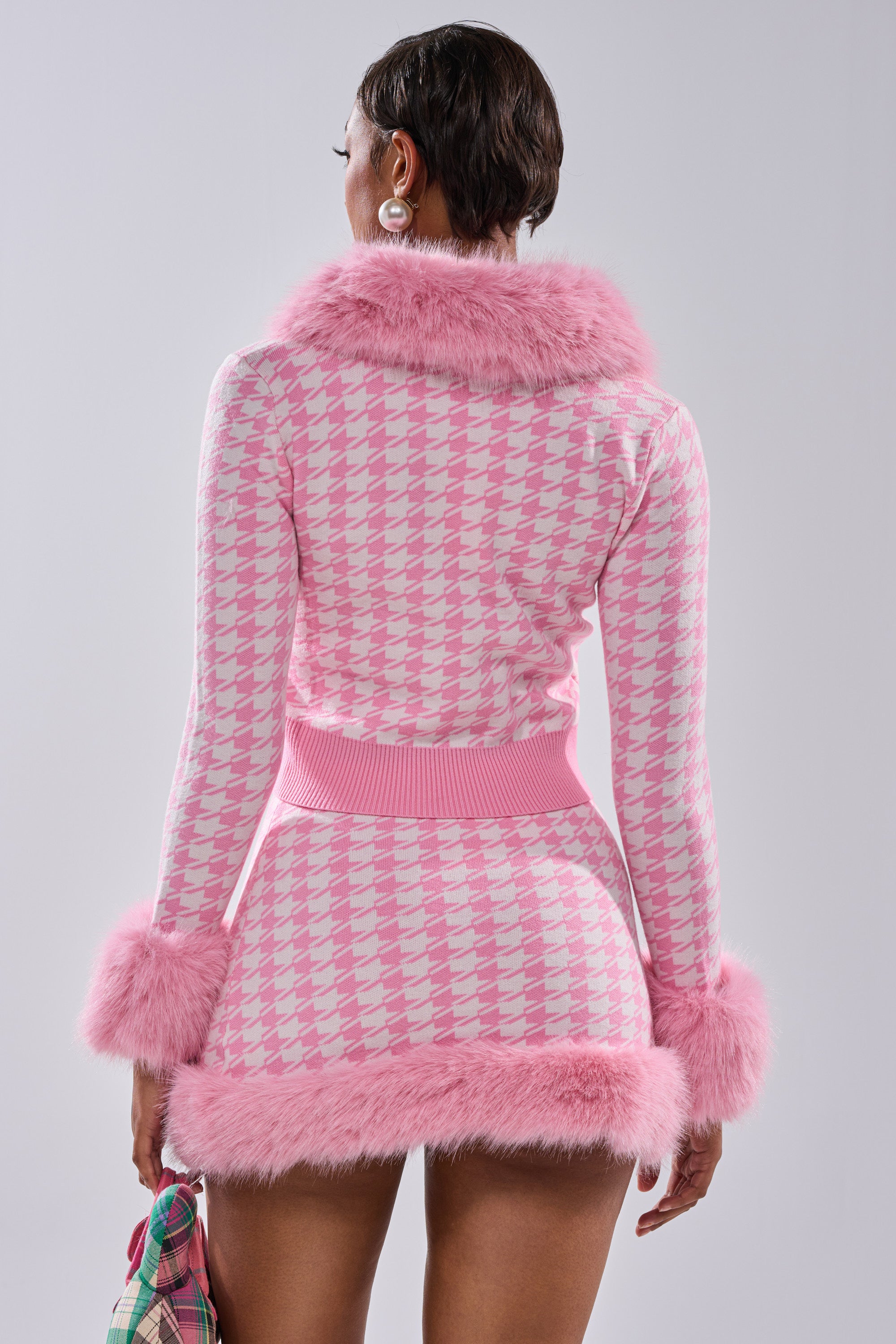 Wearing the QUEEN OF LOVE SWEATER WITH FUR TRIM, a person faces away, showing off its pink and white houndstooth knit and fluffy pink fur accents. They pair it with a bag, pearl earrings, and a chic, short hairstyle.