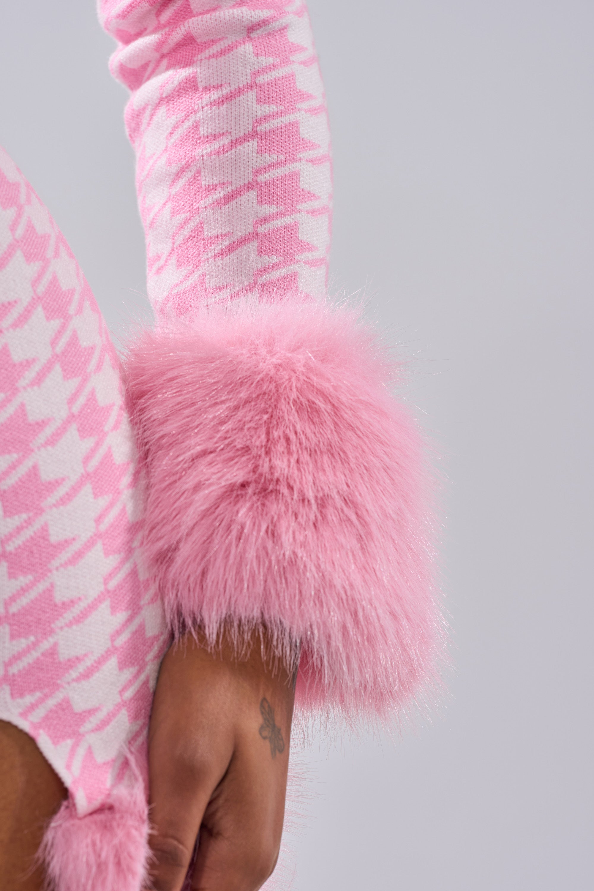 Wearing the QUEEN OF LOVE SWEATER WITH FUR TRIM in bubblegum pink houndstooth and plush faux fur cuffs, a person rests their tattooed wrist at their side against a plain, light background.