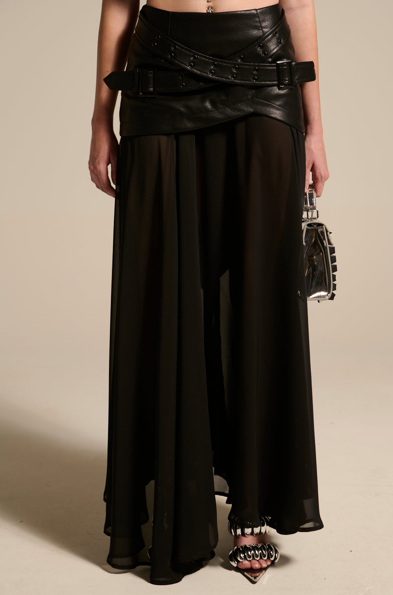 A person wears the DARK NIGHT MIDI SKIRT—a flowing chiffon midi with high-rise waistband and panel details—paired with a small boxy handbag and pointed-toe heeled shoes with metallic embellishments.