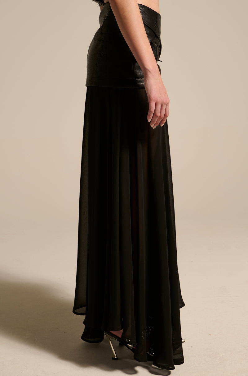 A person stands in profile against a plain beige background, wearing the DARK NIGHT MIDI SKIRT—a black faux leather mini skirt layered over a sheer, floor-length black skirt with a high-rise waistband—and black high-heeled shoes.