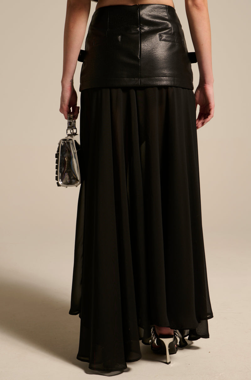 A woman faces away, wearing the DARK NIGHT MIDI SKIRT featuring a faux leather mini over a sheer black maxi design with a high-rise waistband. She holds a metallic handbag and pairs the look with silver high heels against a neutral backdrop.