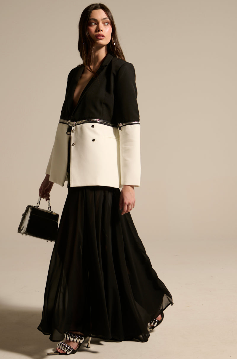 A woman wears the DARK NIGHT MIDI SKIRT—a black, high-waisted, floor-length skirt—paired with a black and white zippered blazer. She accessorizes with a small black handbag and embellished open-toe heels, posing against a beige background.