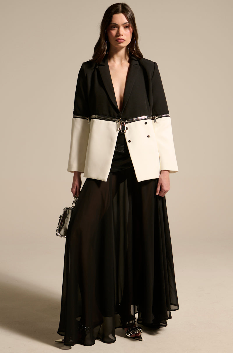 A woman with long brown hair and a neutral expression stands against a plain background, wearing the DARK NIGHT MIDI SKIRT paired with a black and white blazer with silver zipper detail, and holding a small handbag.