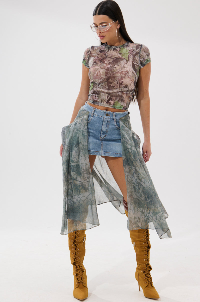 A woman wearing a sheer patterned top, the BRENDA DENIM MINI SKIRT WITH SIDE TIES, a semi-transparent shawl around her waist, yellow lace-up knee-high boots, and glasses stands against a plain white background.