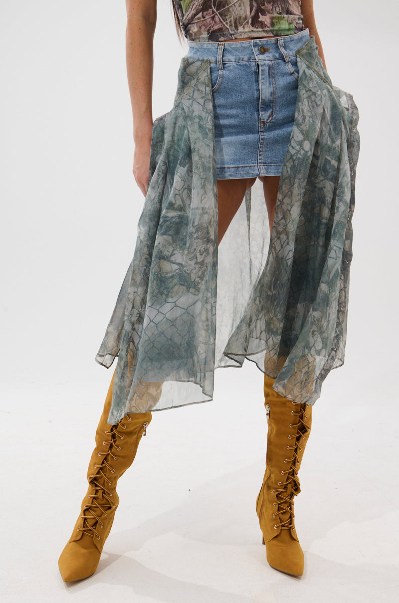 A person wears the BRENDA DENIM MINI SKIRT WITH SIDE TIES featuring sheer mesh panels and a patterned overlay, paired with mustard yellow lace-up knee-high boots, standing against a plain white background.