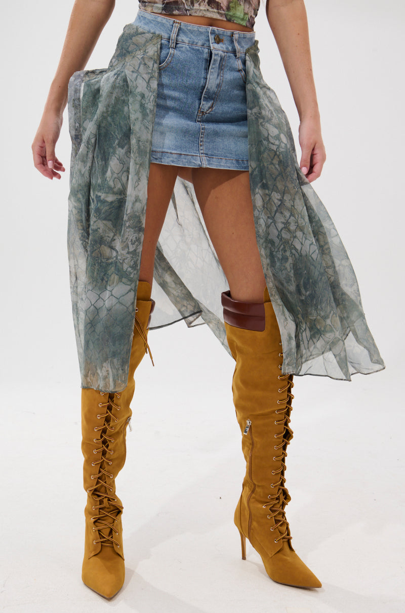 Wearing the BRENDA DENIM MINI SKIRT WITH SIDE TIES, a sheer green patterned overlay, and tall mustard-yellow lace-up heeled boots, a person stands against a plain light background.