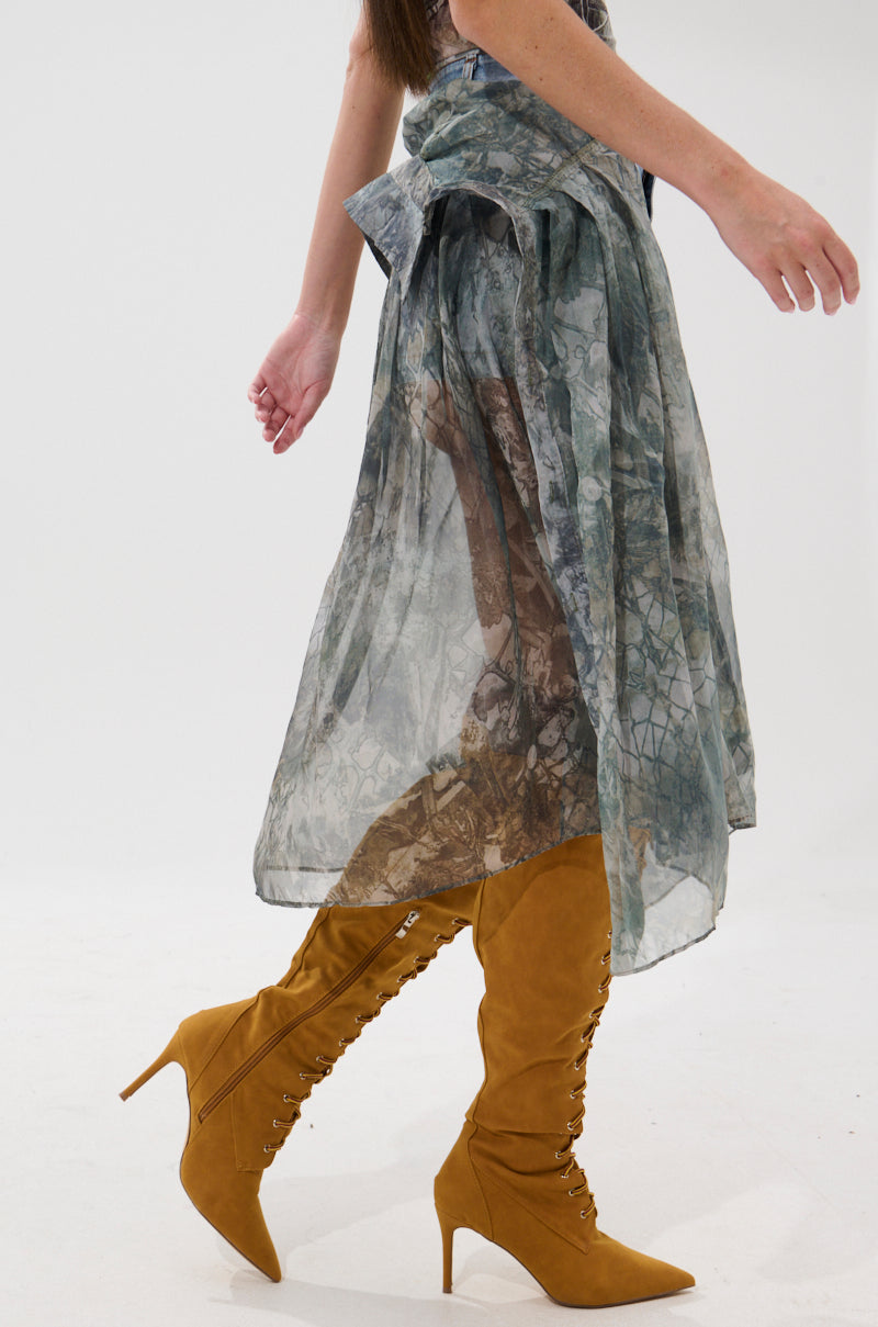 A person wearing the BRENDA DENIM MINI SKIRT WITH SIDE TIES paired with mustard yellow lace-up high-heeled boots, walking against a plain white background.
