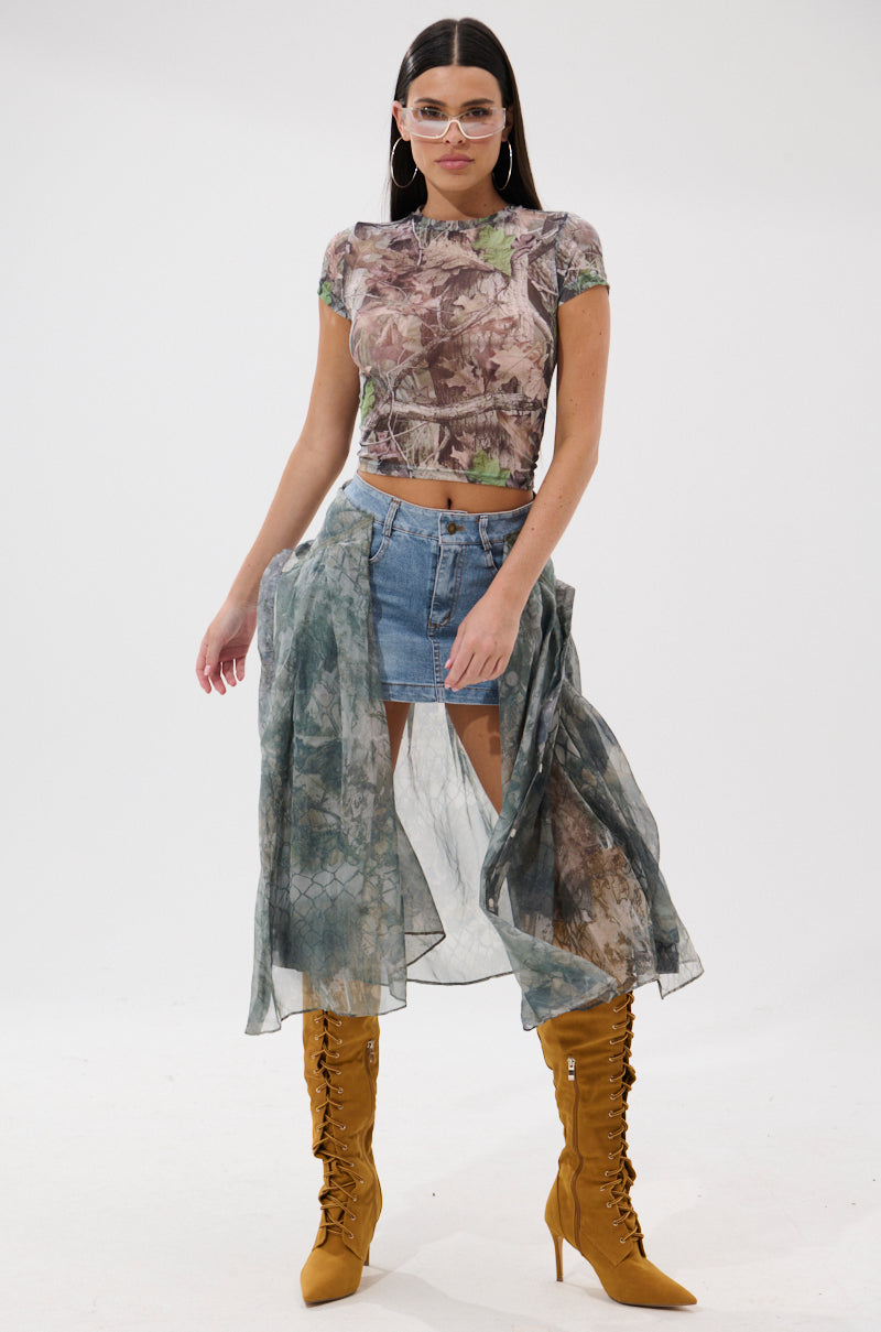 A woman wearing clear glasses, a camo-print crop top, a sheer flowy overlay, knee-high yellow lace-up boots, and the BRENDA DENIM MINI SKIRT WITH SIDE TIES stands against a plain white background.