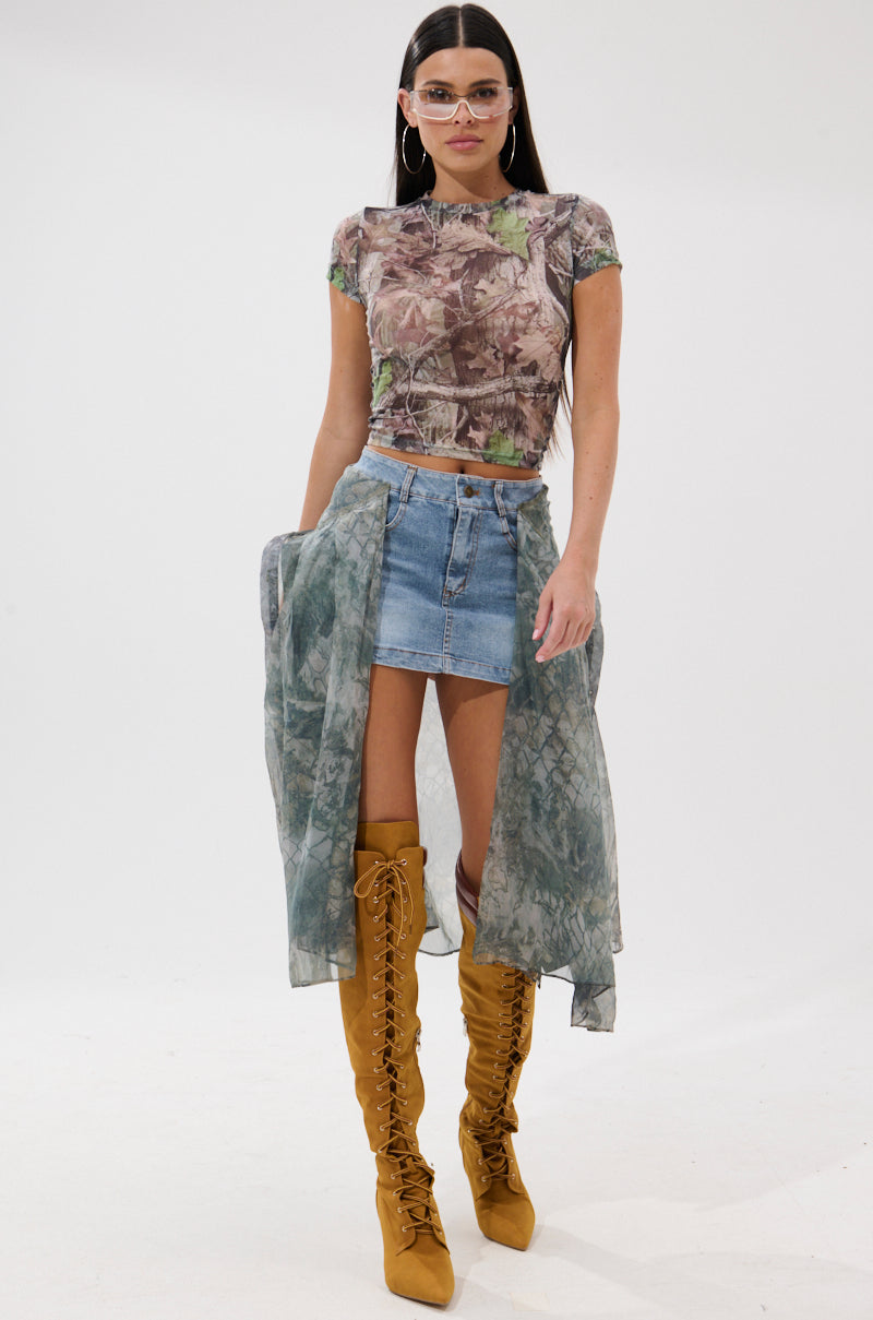 A woman with long dark hair wears clear glasses, a sheer patterned crop top, the BRENDA DENIM MINI SKIRT WITH SIDE TIES, a flowing tie-dye wrap, and mustard yellow lace-up knee-high boots against a plain white background.