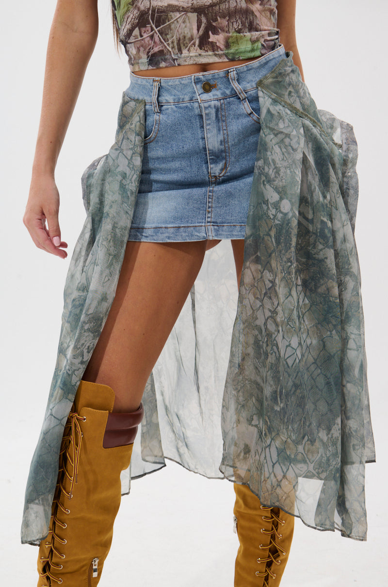 A person wearing the BRENDA DENIM MINI SKIRT WITH SIDE TIES, a sheer green patterned overlay, a camouflage-style top, and tan knee-high lace-up boots. The image is cropped from chest to knees.