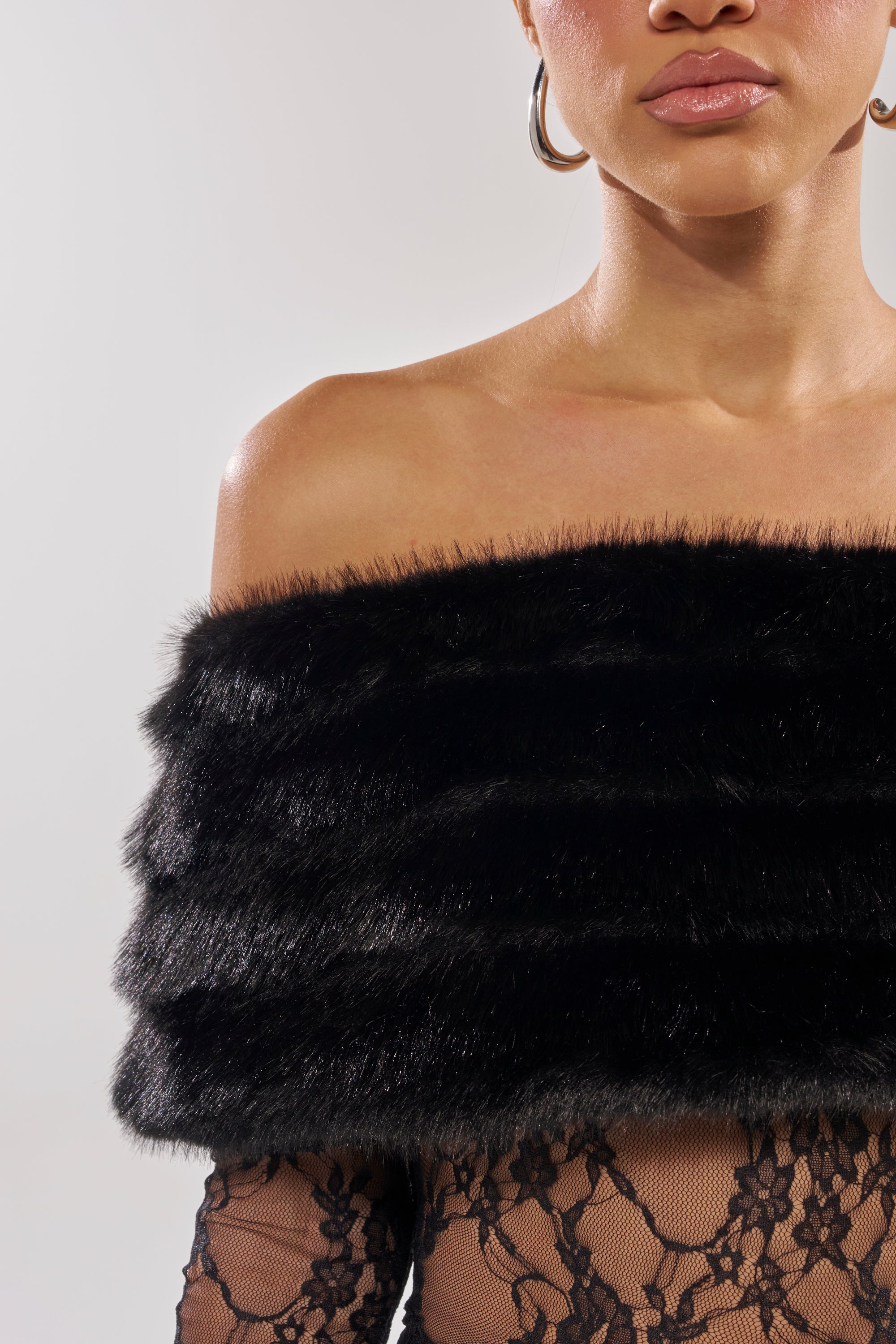 A woman models the LACE N FUR SLEEVELESS JUMPSUIT and a black faux fur off-the-shoulder shawl with large hoop earrings, shown from shoulders to just above the lips against a plain background. 6