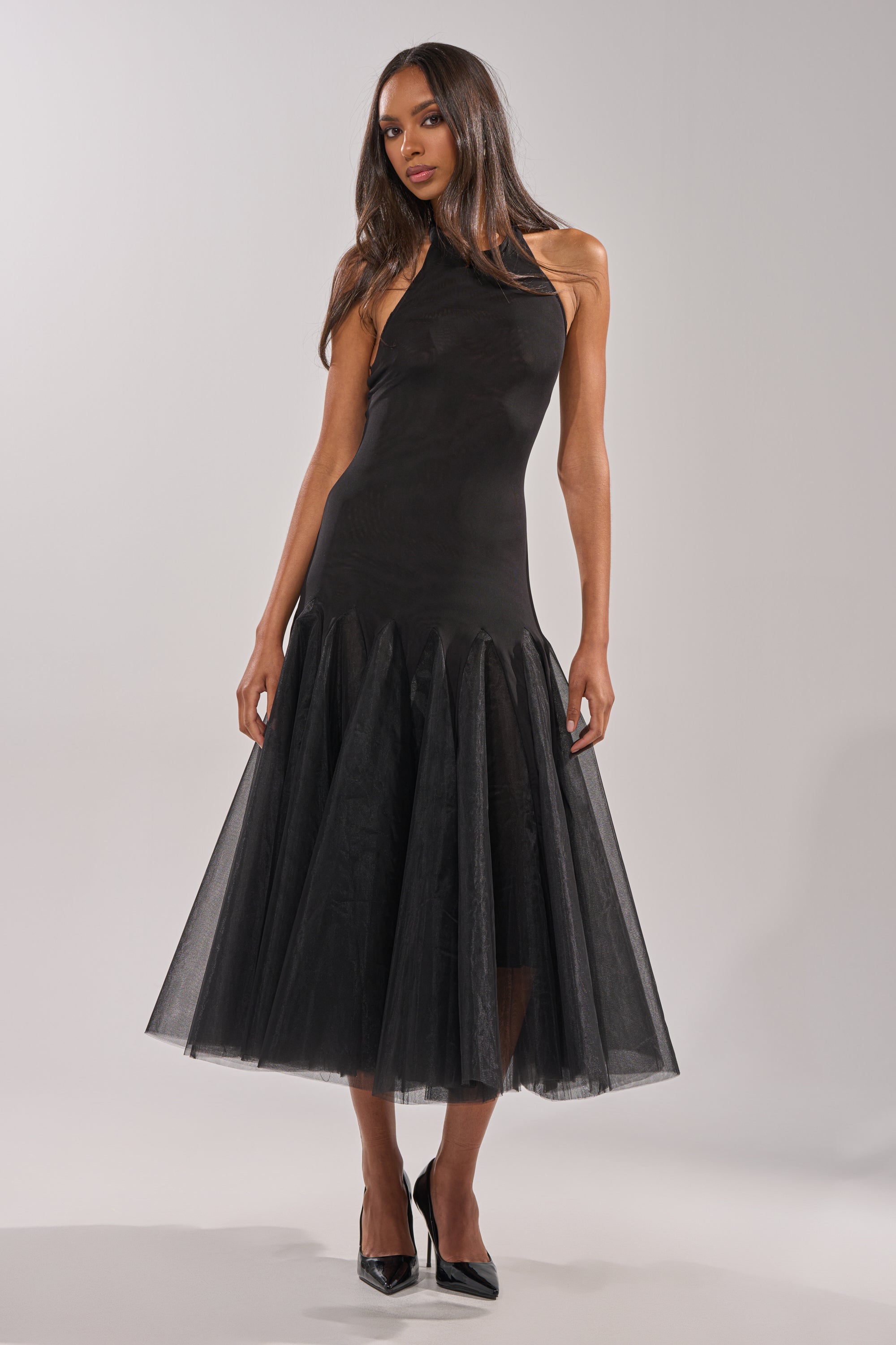 A woman stands against a plain light background in the SWEPT UP MESH MAXI DRESS—a sleeveless black formal gown with a flared, sheer tulle skirt—her long hair down, finished with black pointed-toe heels.