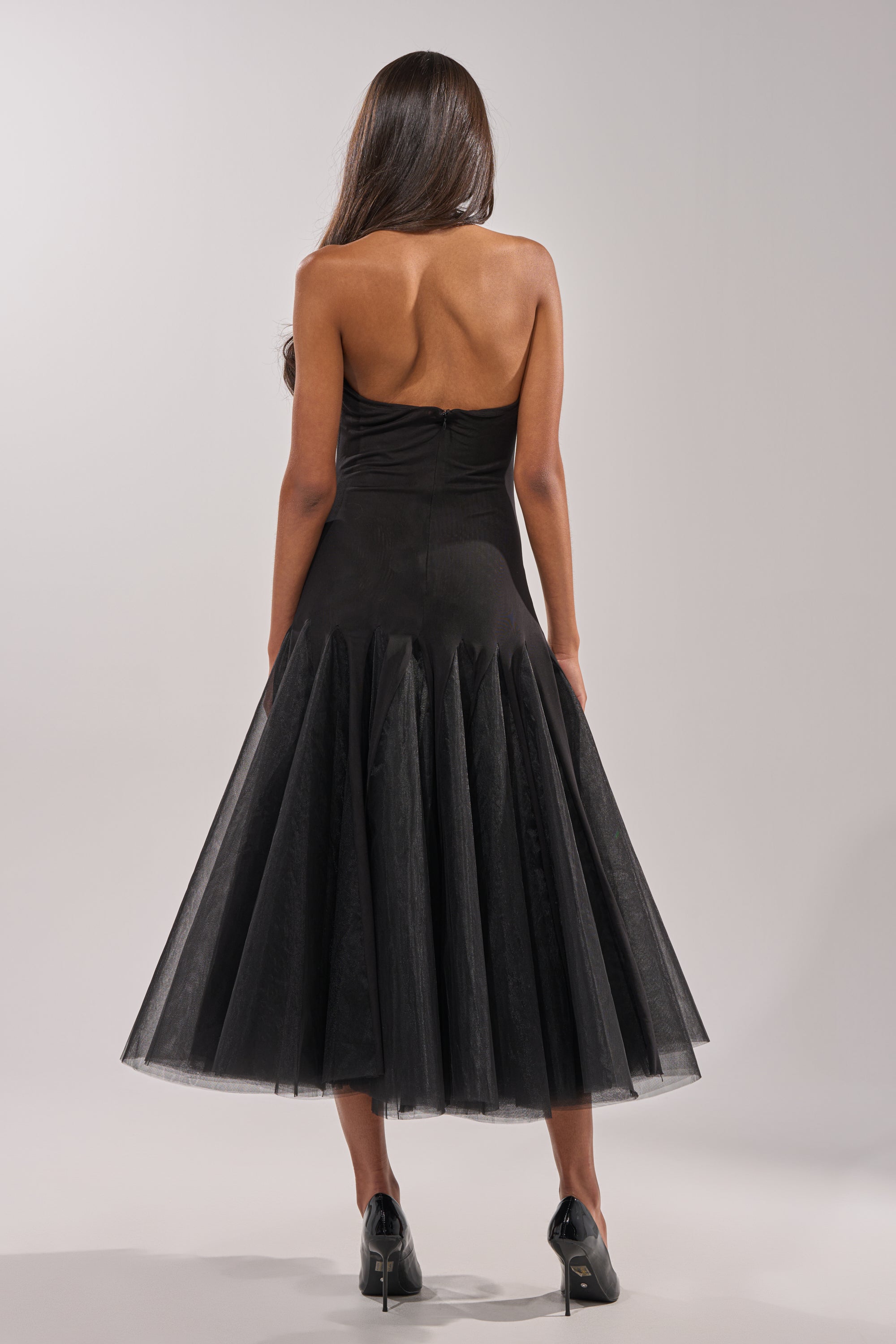 A woman with long brown hair faces away, wearing the SWEPT UP MESH MAXI DRESS featuring a flared tulle mermaid skirt and black high heels, set against a plain light background.