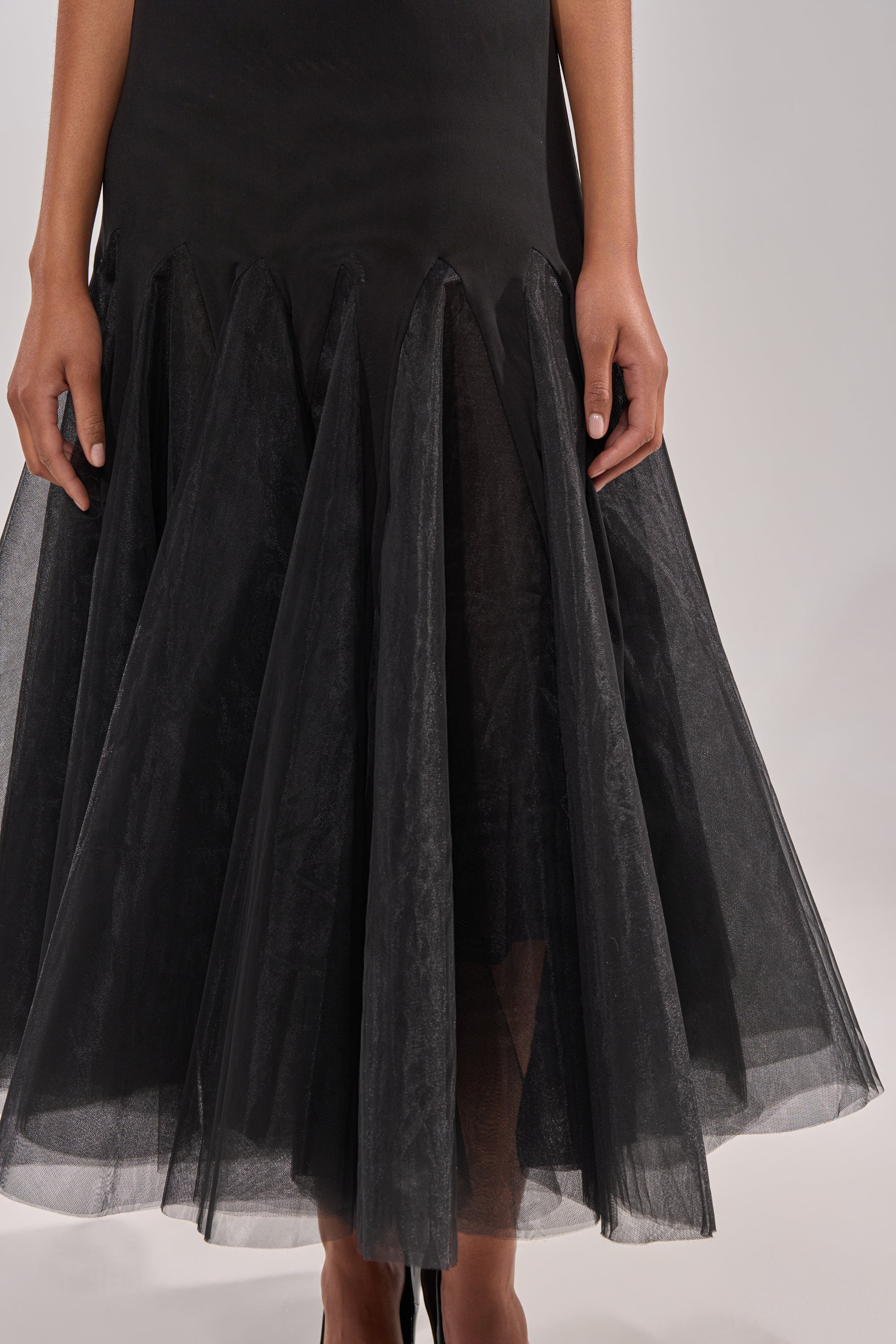 The image shows the lower half of the SWEPT UP MESH MAXI DRESS, a black formal gown with pointed panels and a flared, sheer tulle mermaid skirt. The wearer’s hands rest relaxed by their sides.