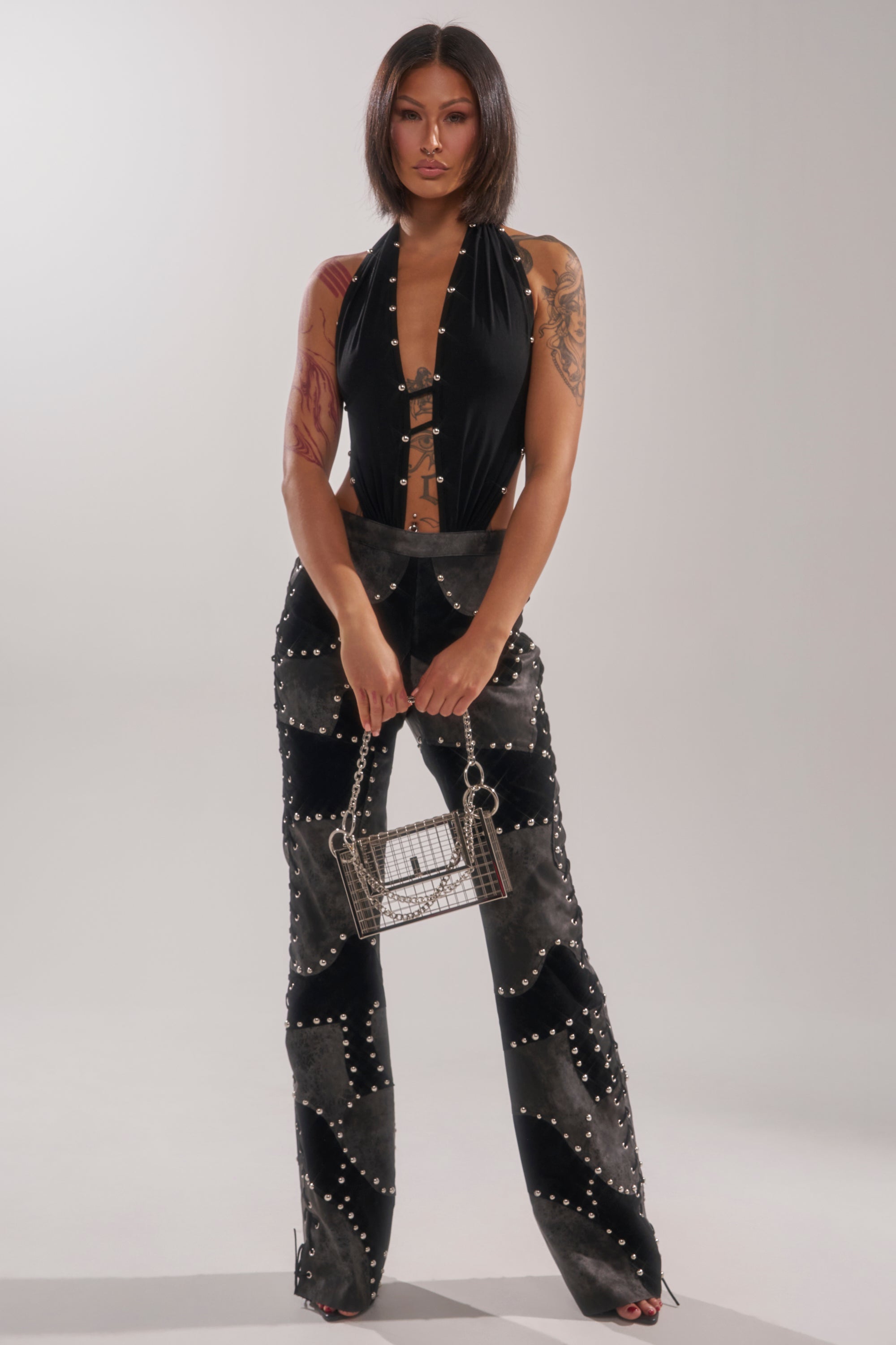 A woman with a bob haircut poses in the ROXY STUDDED SLEEVELESS JUMPSUIT, featuring black studded details and lace-up accents, holding a small silver handbag against a plain light gray background.