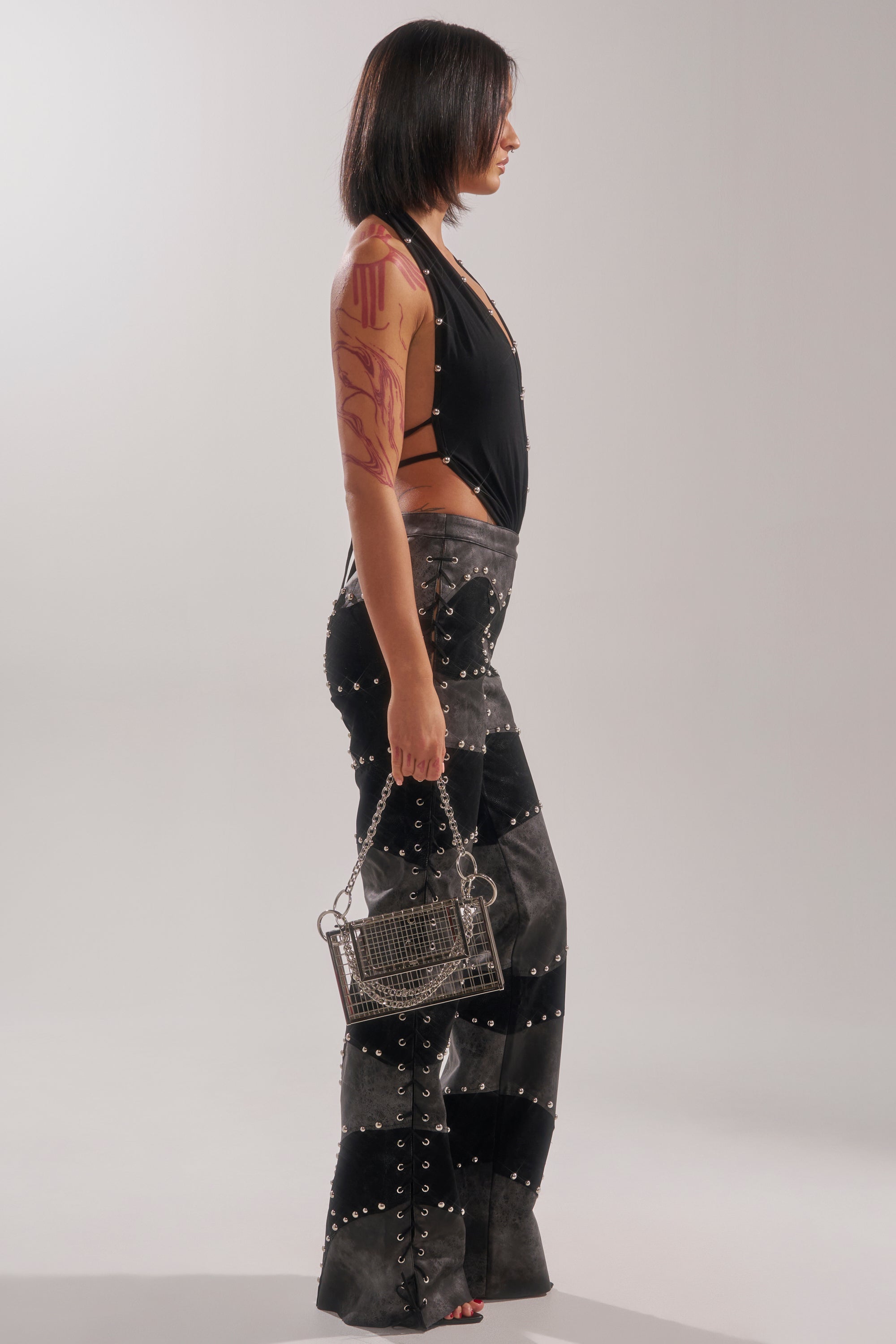 A woman with a visible arm tattoo and shoulder-length dark hair stands in profile, wearing the ROXY STUDDED SLEEVELESS JUMPSUIT with flared legs and holding a small metallic handbag against a plain background.