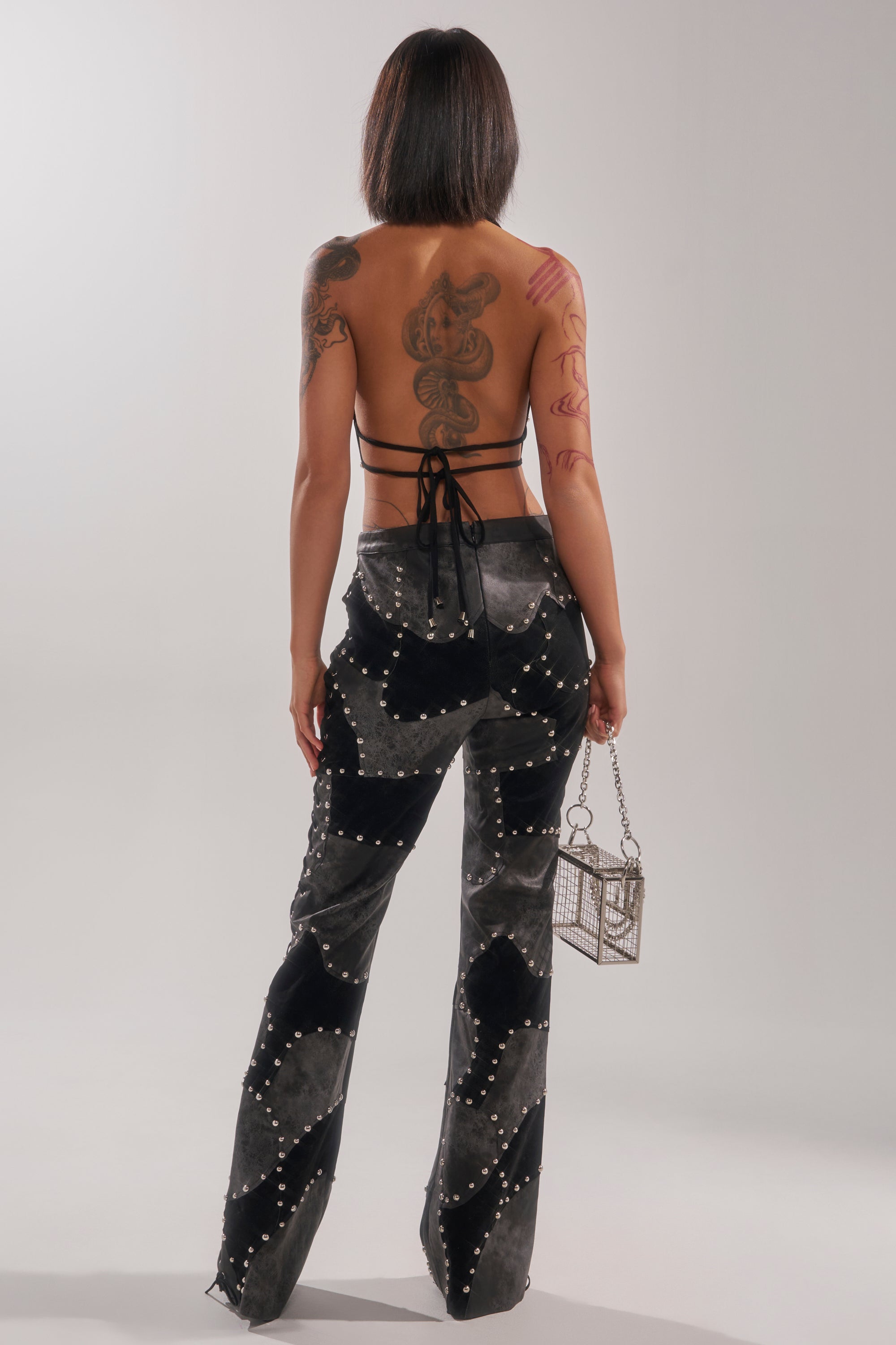 A tattooed woman stands facing away against a plain light background, wearing the ROXY STUDDED SLEEVELESS JUMPSUIT and holding a small box-shaped handbag in her left hand.