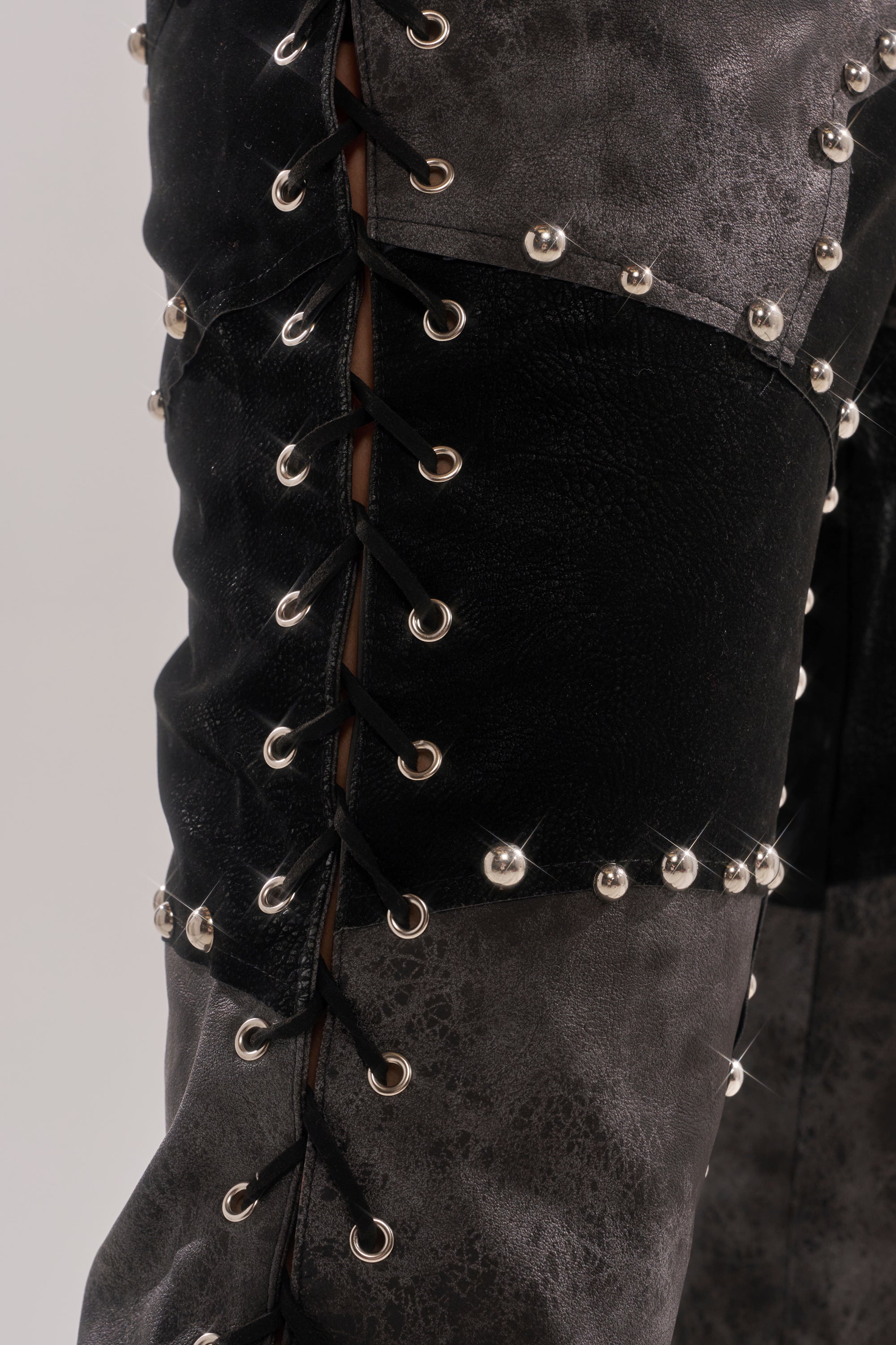 Close-up of the ROXY STUDDED SLEEVELESS JUMPSUIT in black and gray textured faux leather, featuring metal studs, eyelet lace-up details, and an edgy punk-inspired design.