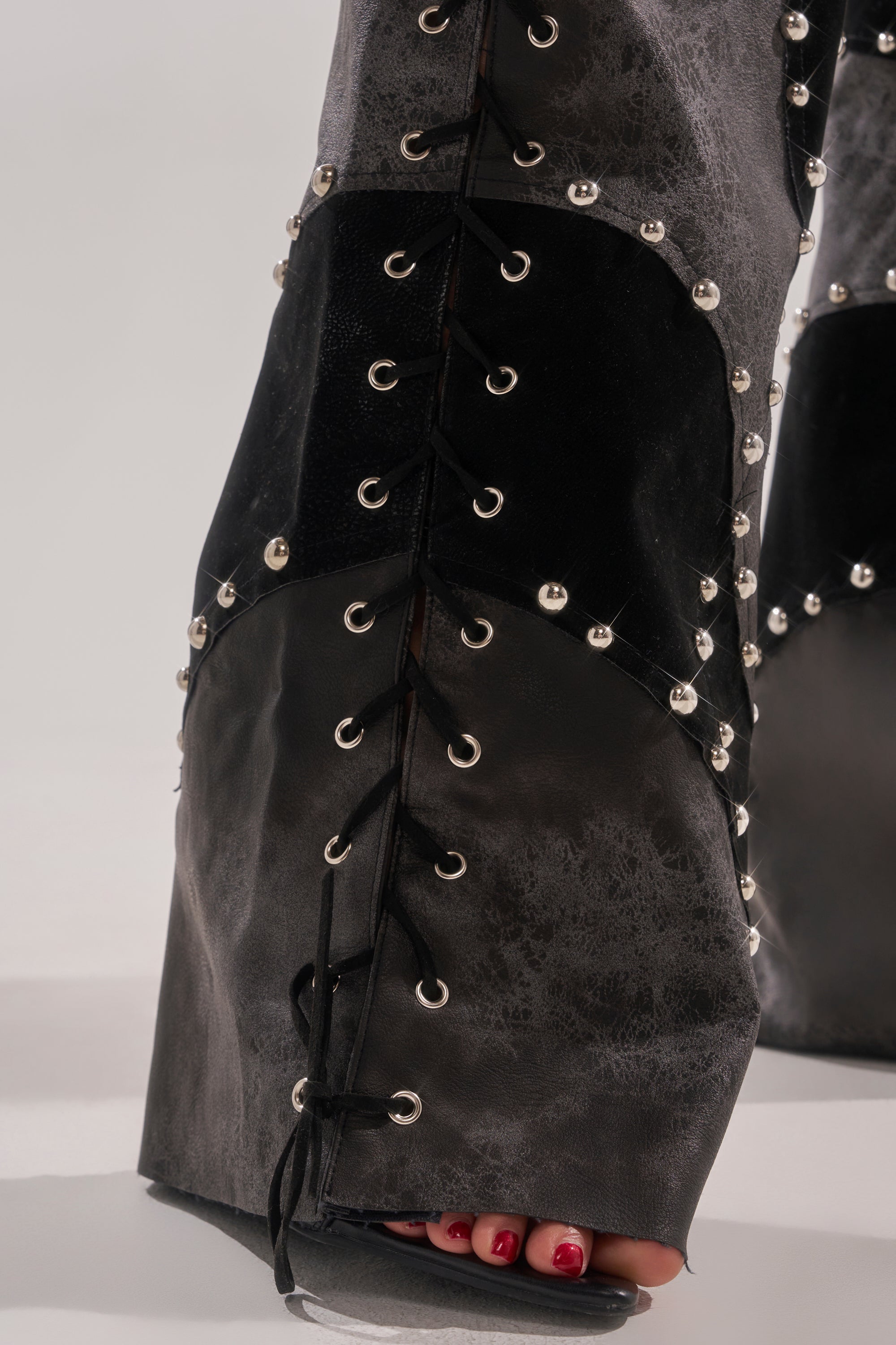Close-up of the ROXY STUDDED SLEEVELESS JUMPSUIT featuring lace-up detailing, multiple textured panels, and silver studs. Red-painted toenails peek through open-toe footwear. The background is plain and light-colored.