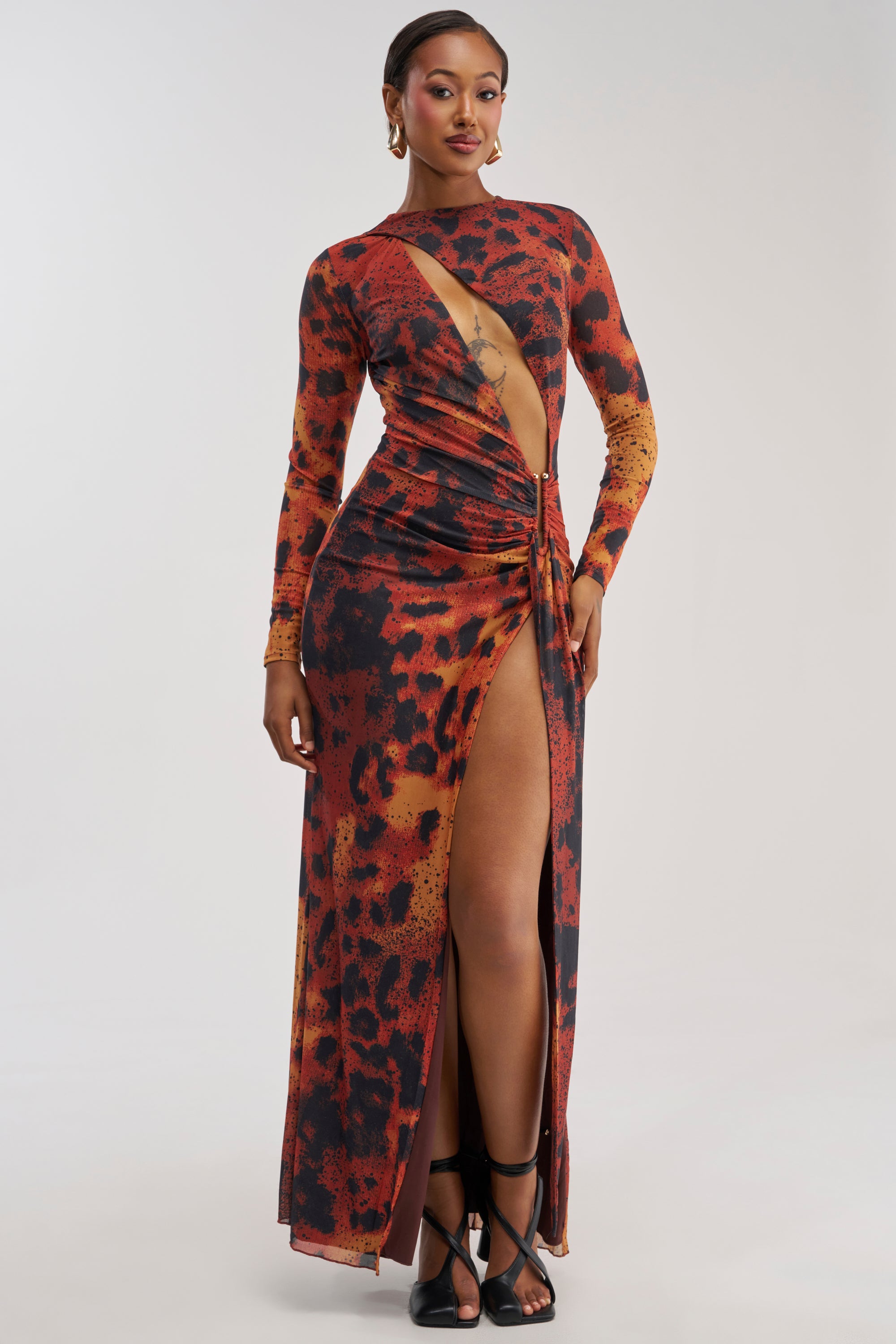 A woman poses confidently in the HOTTEST TOPIC MAXI DRESS IN RED MULTI—a sexy, long-sleeve, red and black abstract print maxi dress featuring a high slit and front cutout—styled with strappy black heels against a plain background.