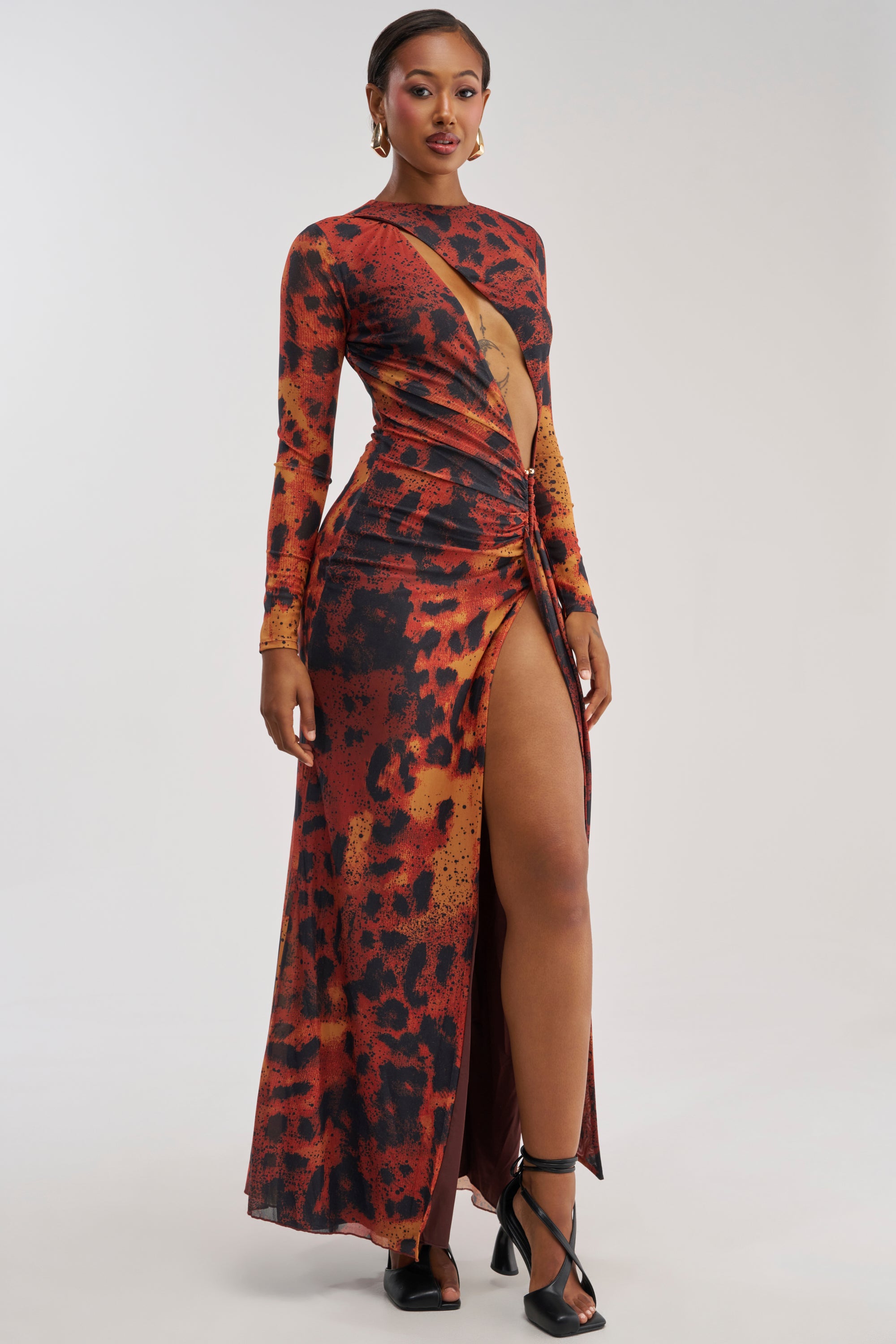 A woman wearing the HOTTEST TOPIC MAXI DRESS IN RED MULTI, a long, fitted maxi dress featuring an abstract red multi print, bold cutout front, high slit, and a striking silhouette, stands in black heels against a plain background—an effortlessly sexy date night look.