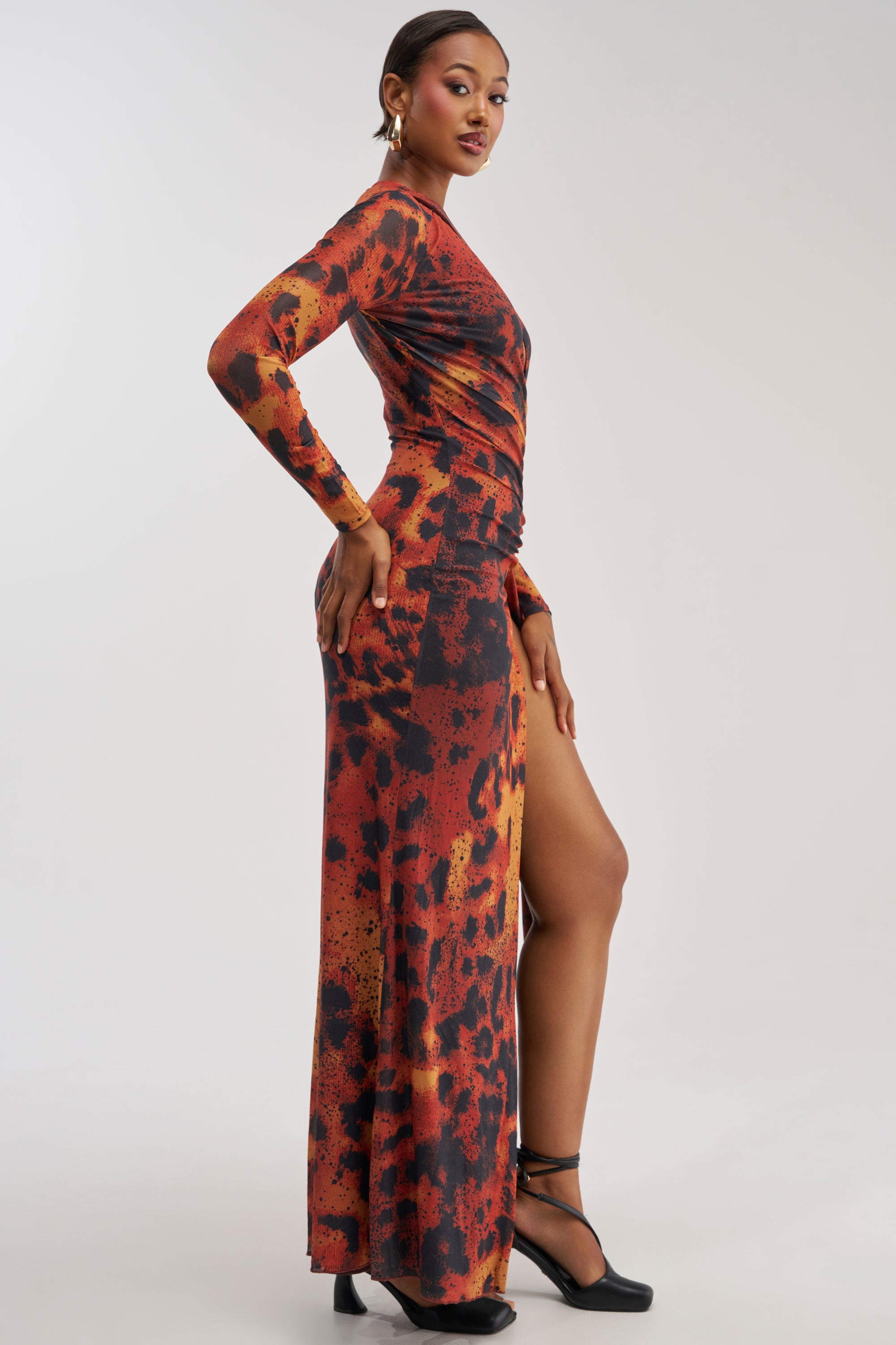 A woman stands sideways against a plain background, wearing the HOTTEST TOPIC MAXI DRESS IN RED MULTI—a long-sleeved, abstract print maxi dress featuring a high slit—styled with black heels and hoop earrings.