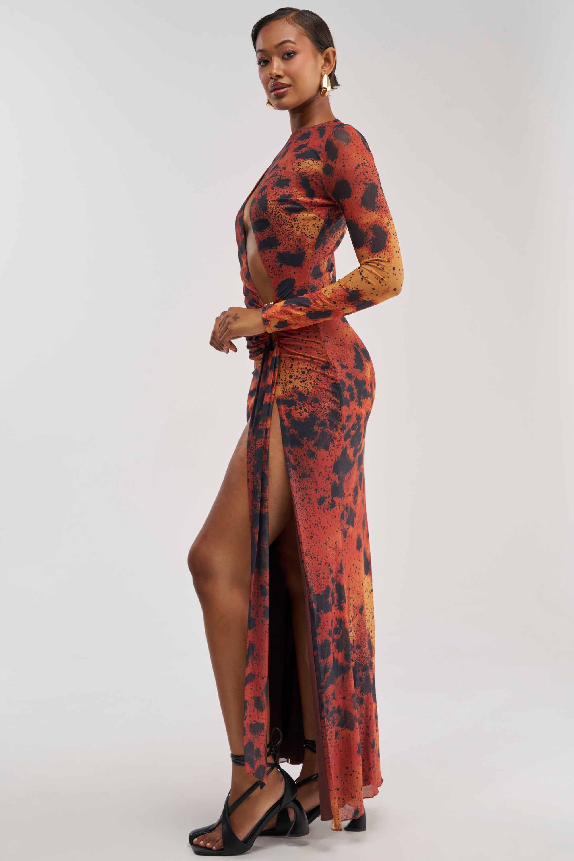 A woman poses in the HOTTEST TOPIC MAXI DRESS IN RED MULTI, a long-sleeve, abstract print maxi dress with a high slit. She completes the sexy date night look with black heeled sandals and gold hoop earrings, standing against a plain white background.
