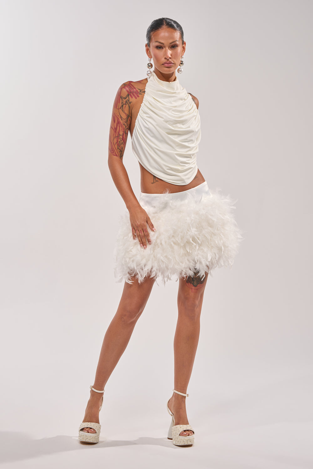 A person stands confidently in the LITTLE FAIRY FEATHER MINI SKIRT paired with a sleeveless white top, white platform heels, large earrings, and visible arm tattoos. 1