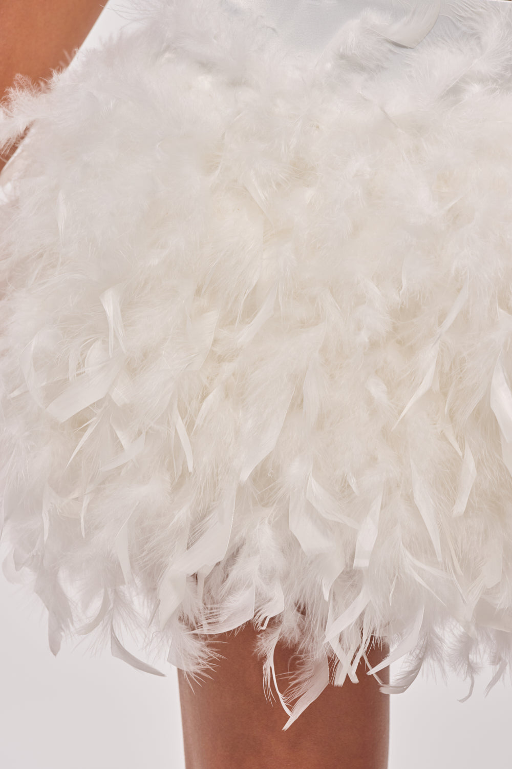 A close-up of a person wearing the LITTLE FAIRY FEATHER MINI SKIRT, showcasing its fluffy feather trim, with their lower arm and leg visible against a plain background. 10