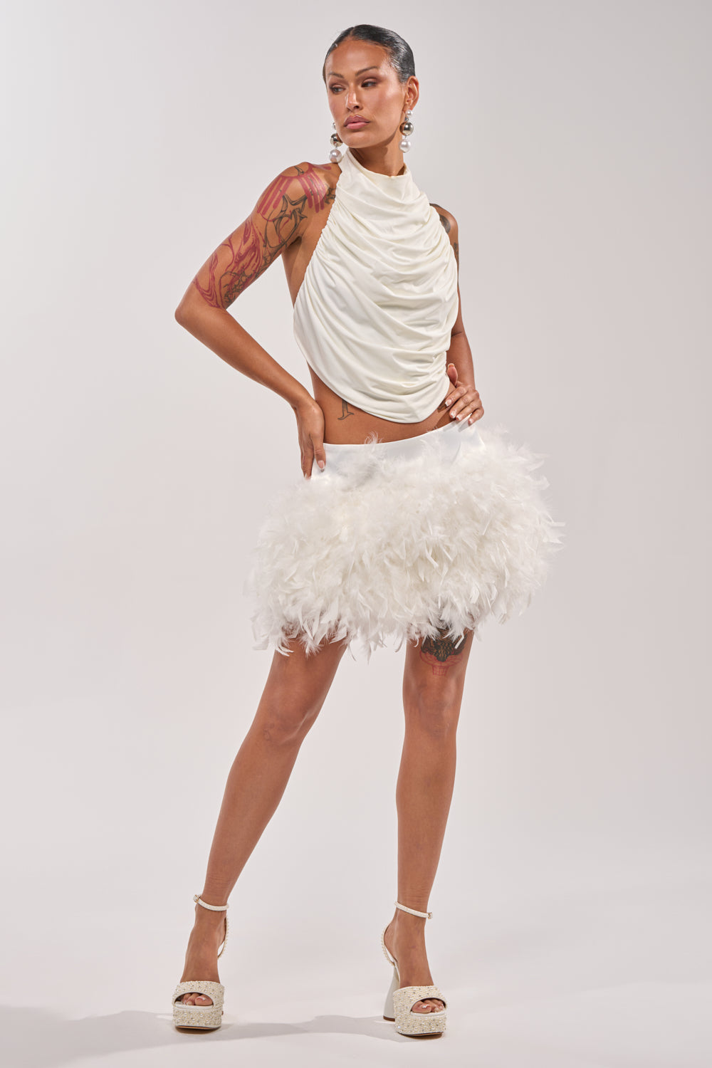 A woman models the LITTLE FAIRY FEATHER MINI SKIRT with a white sleeveless draped top, platform heels, and gold earrings. She displays a tattoo sleeve on one arm against a plain light background. 9