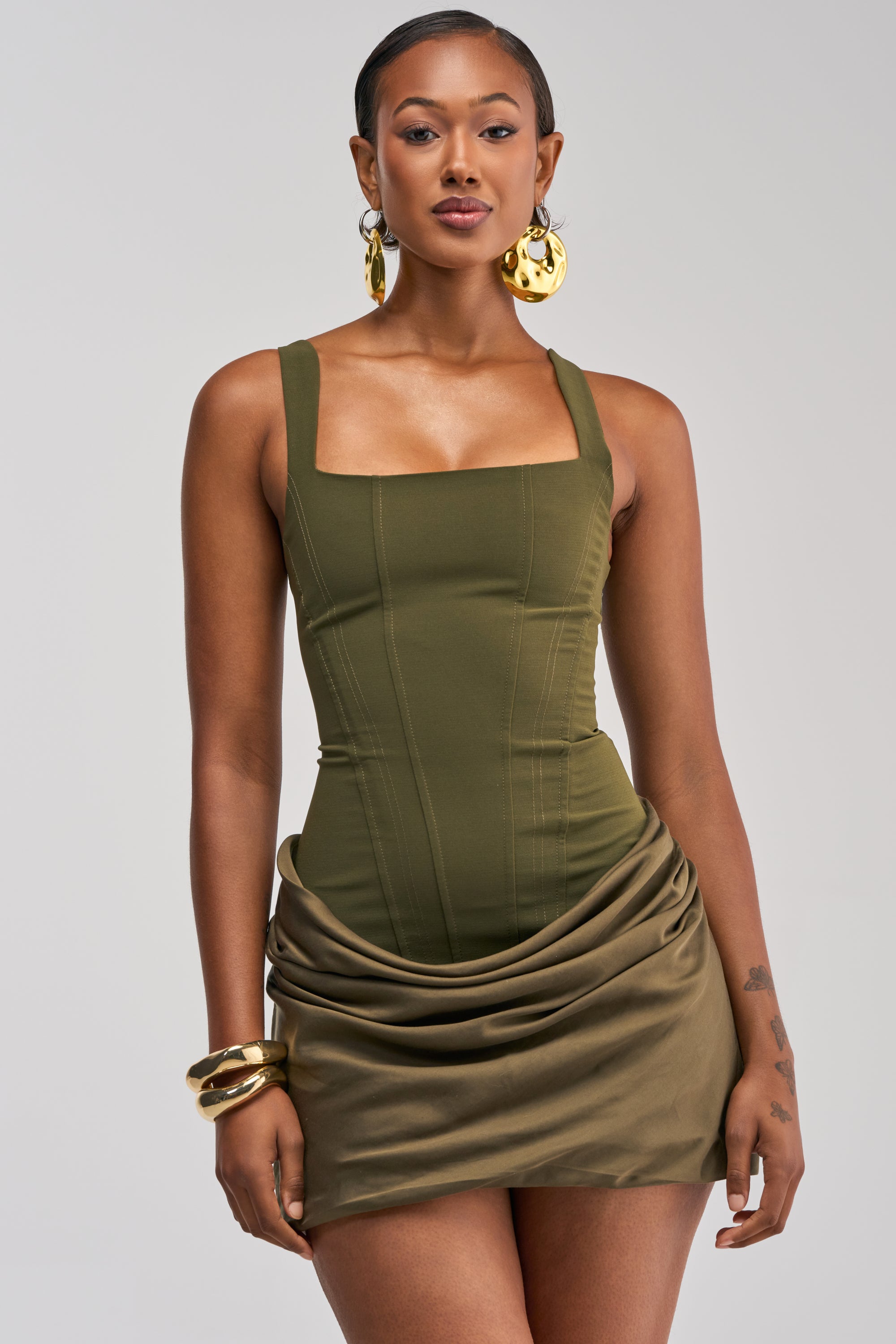 A woman wearing the PLAY NICE CORSET MINI DRESS IN OLIVE—a fitted mini dress featuring a draped satin skirt—styled with large gold hoop earrings and a gold bracelet, stands against a plain light gray background, making this look the perfect choice for a chic date night or an elegant cocktail party.