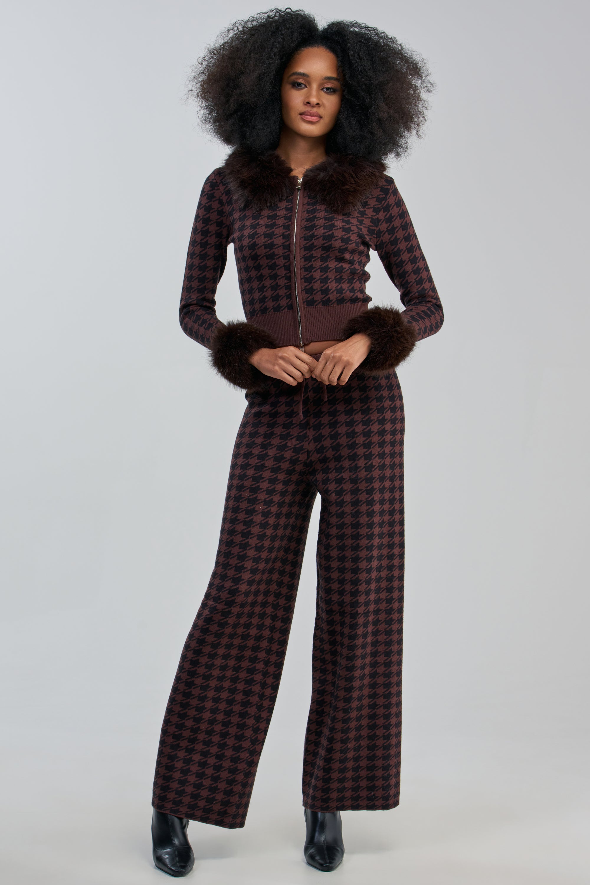 A woman stands with her hands together in front, wearing the HIGH MAINTENANCE HOUNDSTOOTH PANT as part of a brown and black checkered matching set with wide-leg pants and faux fur trim on the cuffs and collar, set against a plain background.