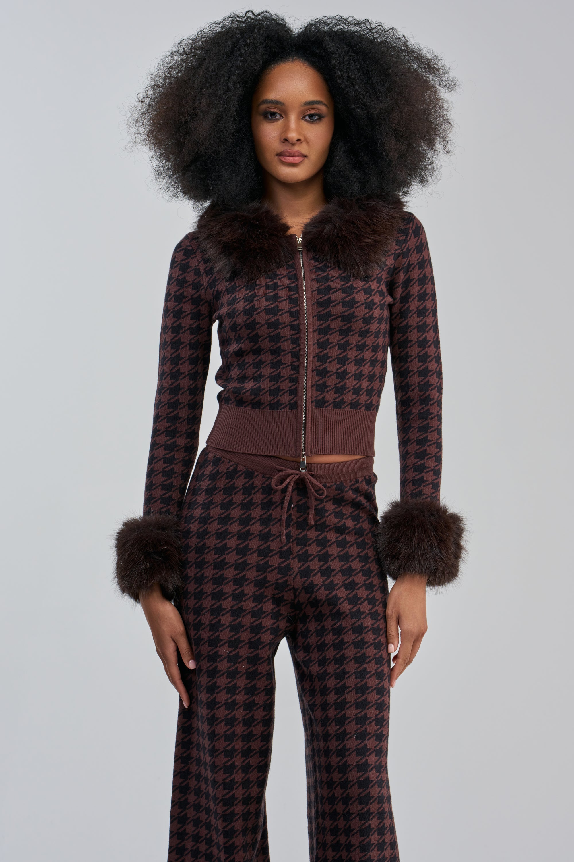 A woman wearing the HIGH MAINTENANCE HOUNDSTOOTH FUR TRIM SWEATER—a brown and black cropped zip-up sweater with a chic houndstooth pattern, accented by faux fur trim on the collar and cuffs—stands against a plain gray background.
