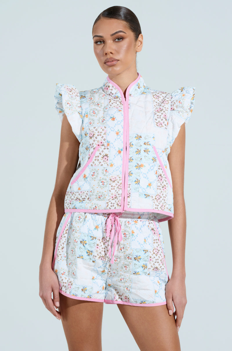 A person with vintage feminine flair stands against a light background showcasing a MIXED FLORAL PUFFER VEST. The outfit features ruffled shoulders, zip-up front, and floral patterns, completed with matching shorts adorned with pink trim.