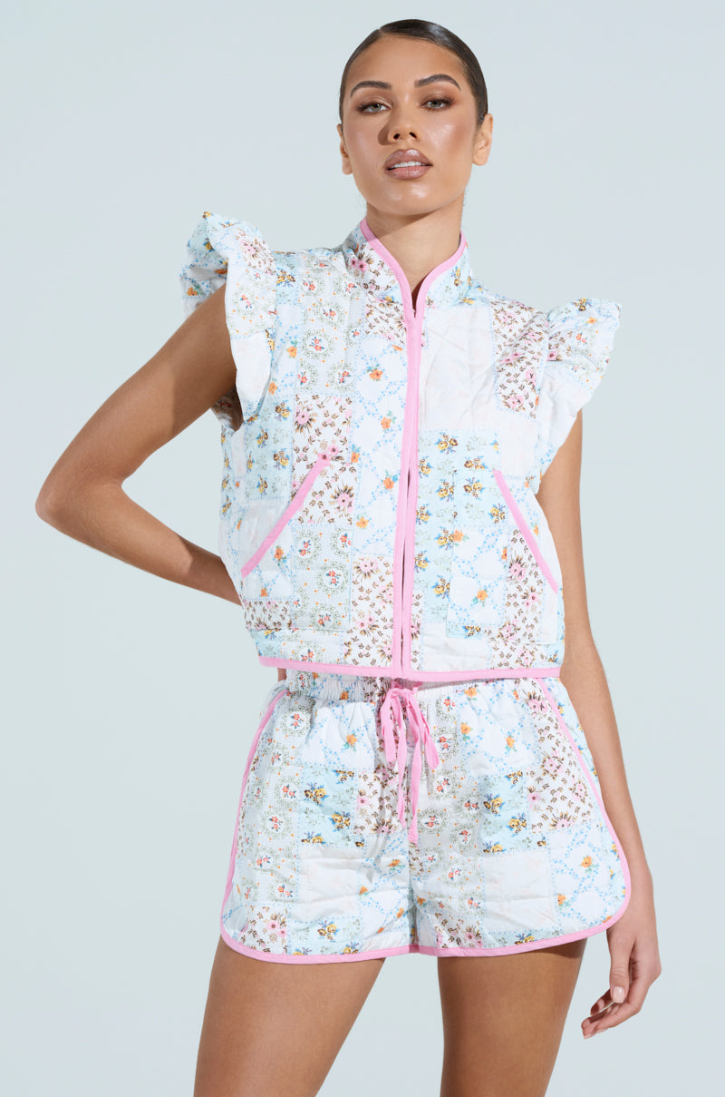 Radiating vintage feminine flair, a person confidently poses hand on hip in the MIXED FLORAL PUFFER VEST, featuring light blue florals with ruffled sleeves and matching shorts, accented by pink trim and drawstring against a plain background.