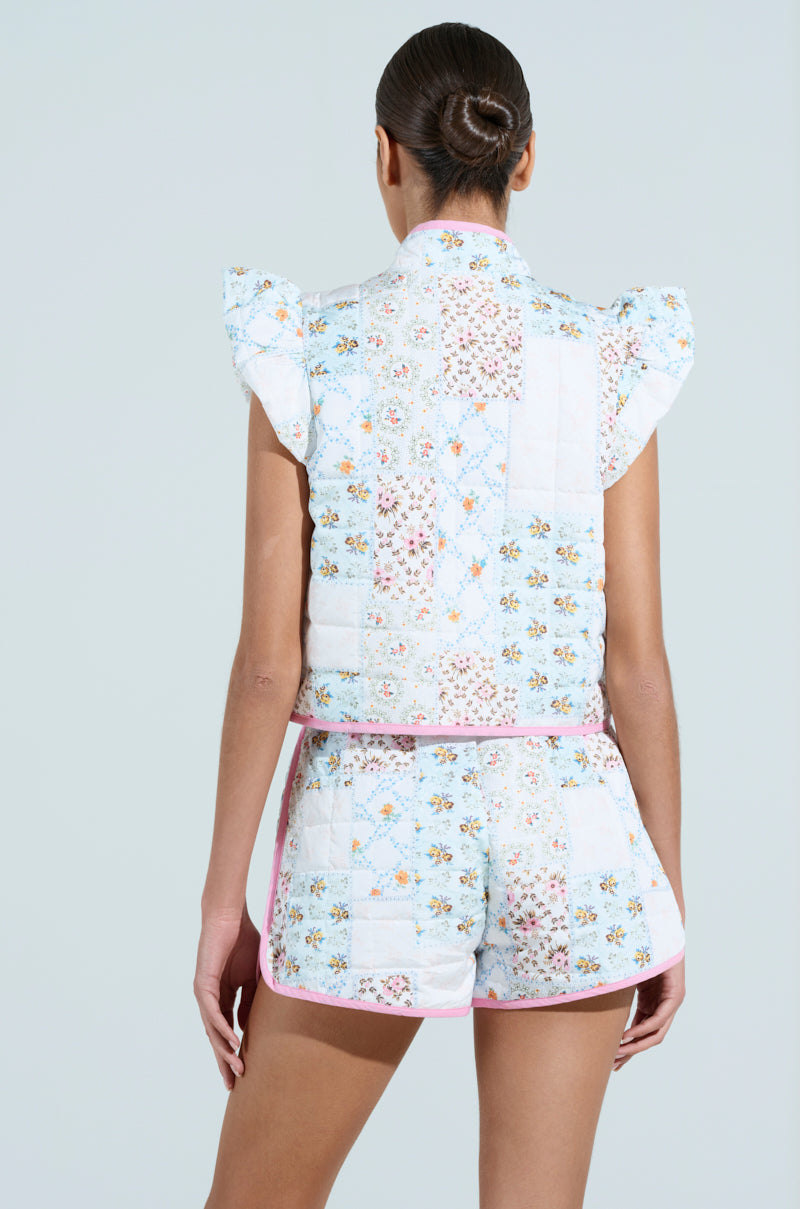 A person with a bun hairstyle is seen from the rear wearing a short-sleeved, floral-patterned quilted top and matching Mixed Floral Puffer Vest with pink trim. The garments exude Vintage Feminine Flair through their patchwork design on a neutral background.