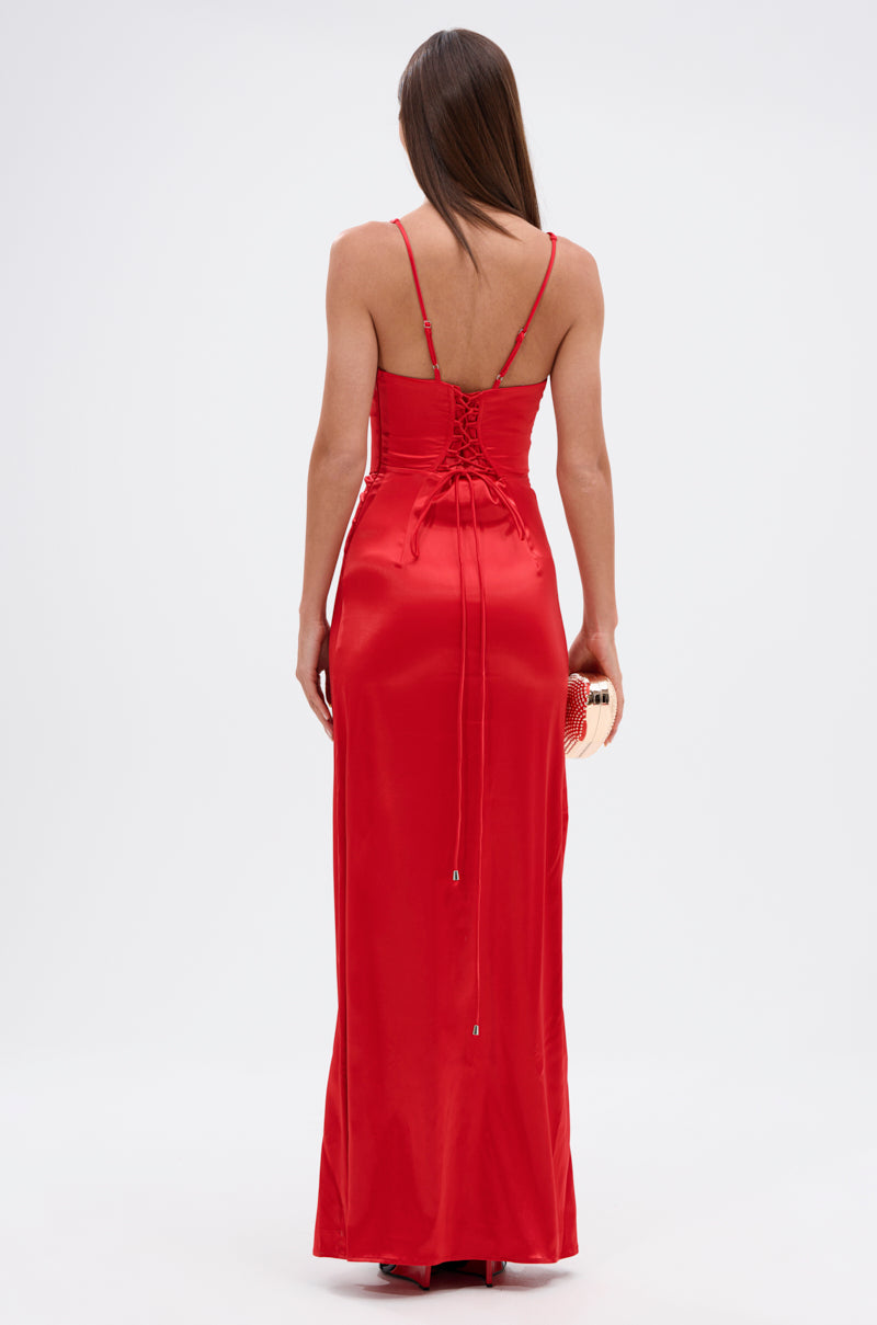 A woman with long straight hair wears the ALL ABOUT ME SATIN MAXI DRESS featuring thin straps and lace-up back detail, standing with her back turned while holding a small clutch purse.