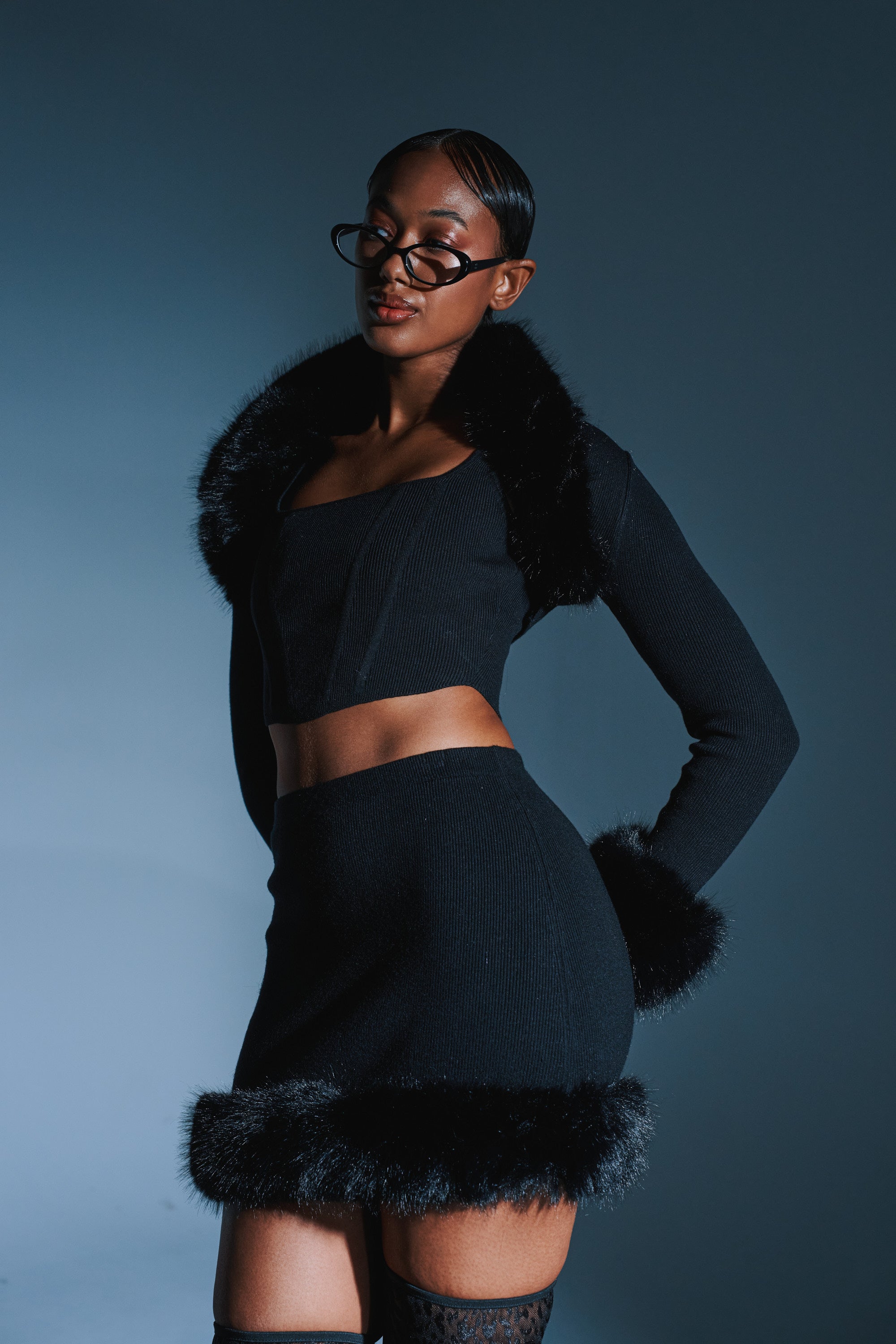 A stylish woman poses confidently in the BIG MOOD FUR TRIM MINI SKIRT IN BLACK, paired with glasses and thigh-high stockings, set against a dark blue background.