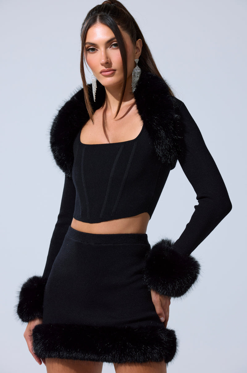 A woman with straight dark hair and statement earrings poses against a plain light background, styled in the BIG MOOD FUR TRIM SHRUG IN BLACK layered over a black crop top and matching mini skirt.