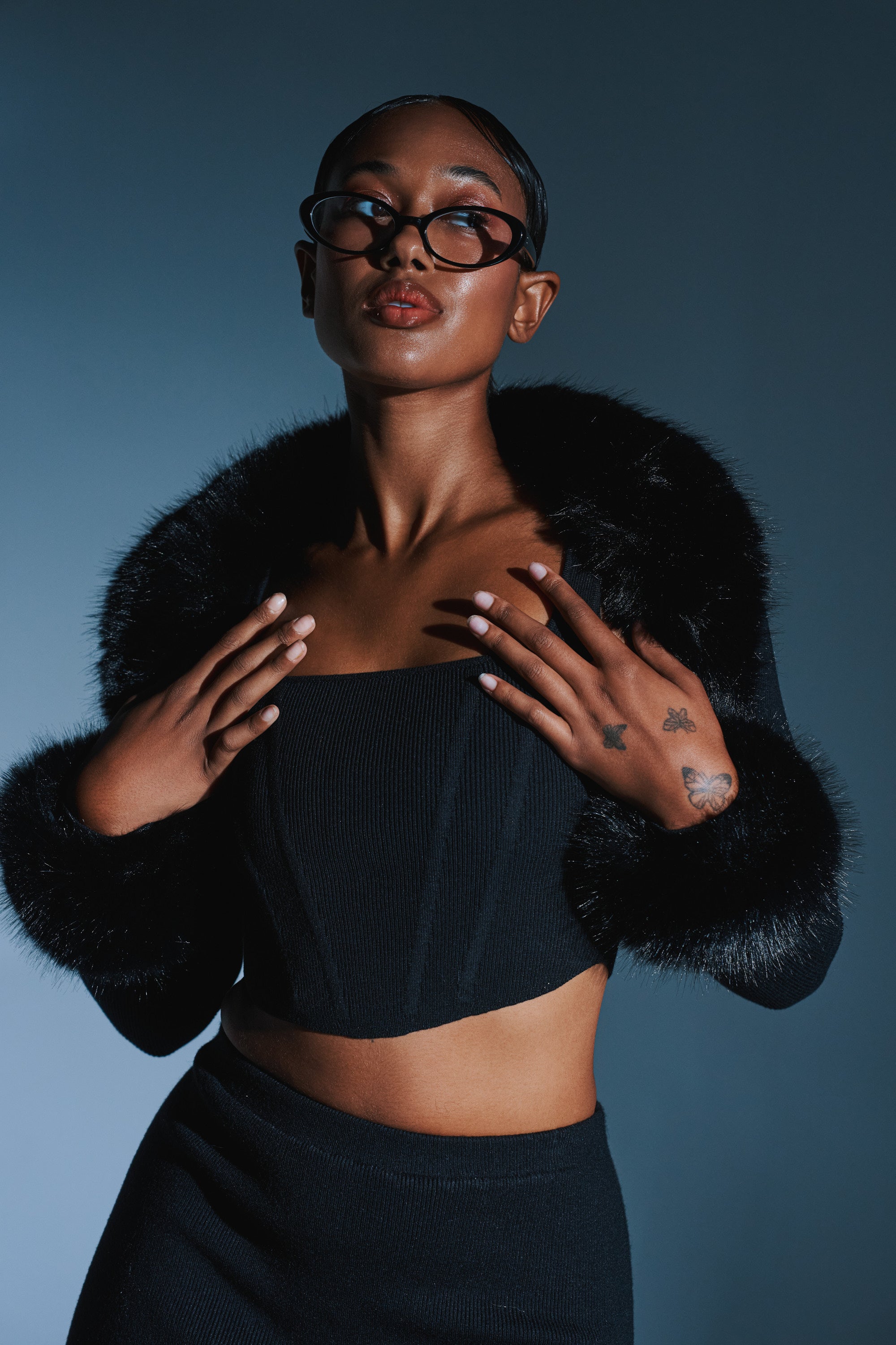A stylish woman with short hair, tattoos, and glossy skin poses confidently against a blue-gray backdrop, wearing the BIG MOOD FUR TRIM SHRUG IN BLACK with a faux fur collar and glasses, hands near her collar.