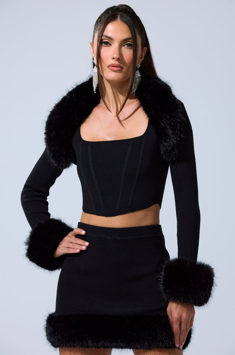 A woman models the BIG MOOD FUR TRIM SHRUG IN BLACK, a chic cropped shrug with black faux fur trim on the collar, cuffs, and hem. She styles it with a crop top, mini skirt, chandelier earrings, and long dark hair.