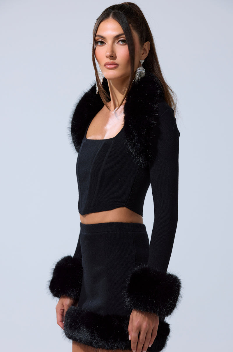 A woman stands against a light gray background wearing the BIG MOOD FUR TRIM SHRUG IN BLACK over a matching black, long-sleeve crop top and mini skirt with faux fur trim for a chic look.