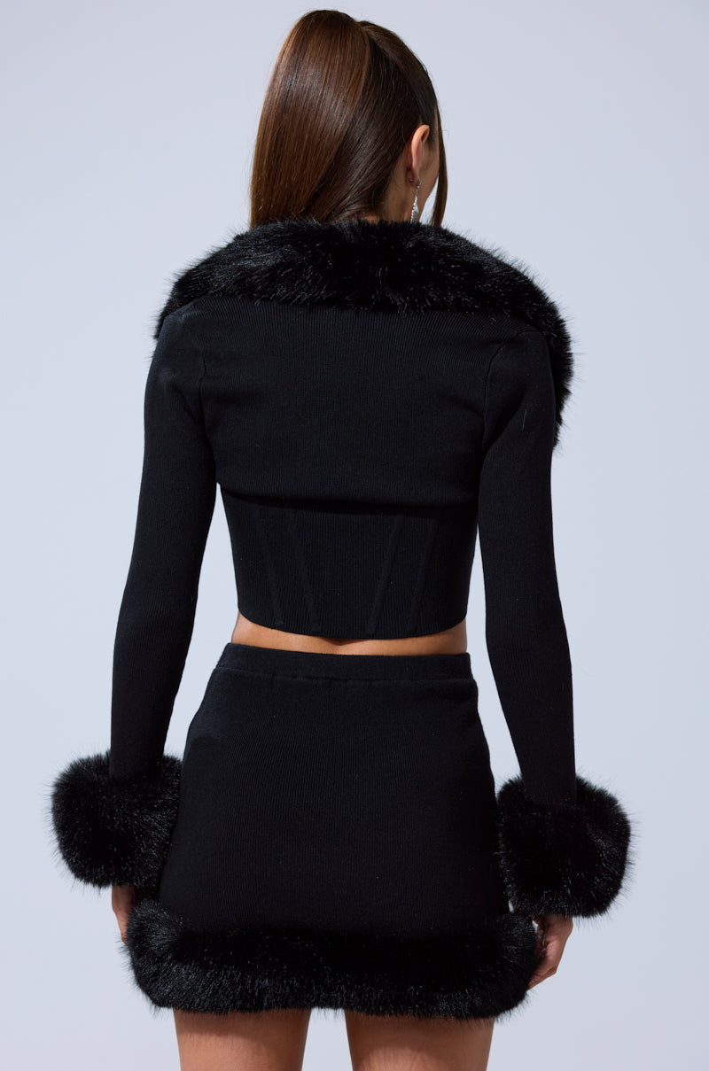 A woman faces away, wearing the BIG MOOD FUR TRIM SHRUG IN BLACK over a matching black long-sleeve crop top and mini skirt with thick faux fur trim; her hair is styled in a high ponytail.