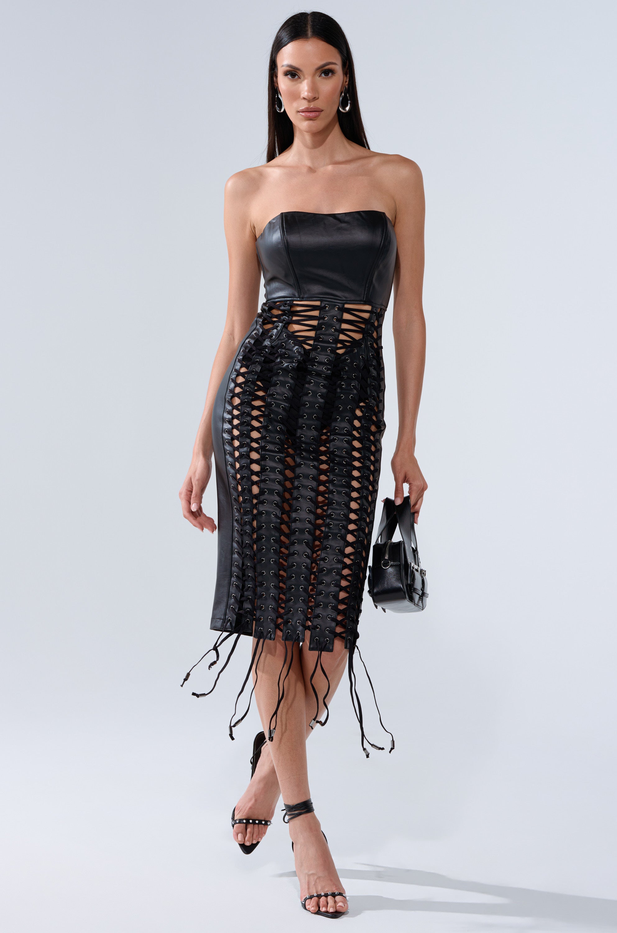 A woman stands confidently in the NOT FOR YOU PU LACE UP MIDI DRESS, featuring a strapless leather bodice and woven skirt, holding a black handbag and wearing strappy heels against a light background.