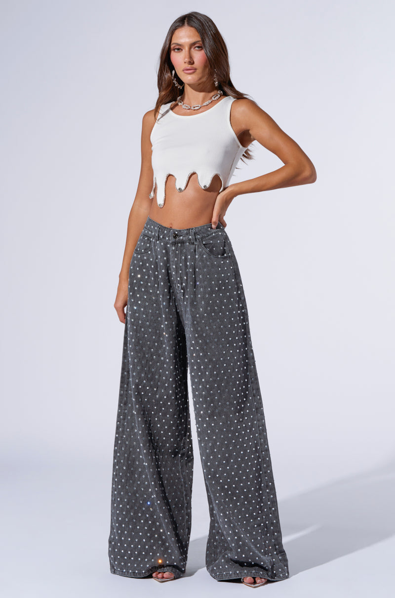 A woman with long hair is wearing a white sleeveless top with a wavy hem and DAZZLED RHINESTONE STUDDED DENIM PANTS IN BLACK. She is standing in a neutral pose against a plain background.