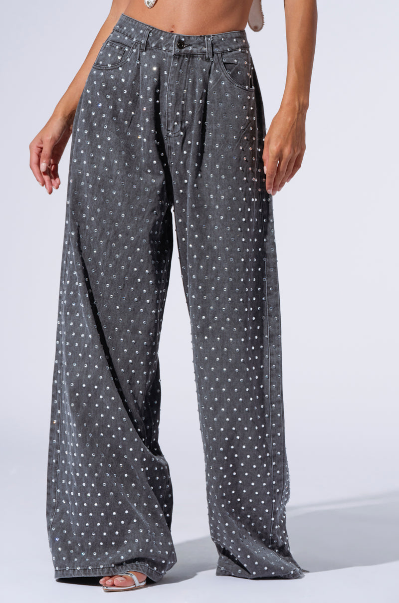 A person models a pair of DAZZLED RHINESTONE STUDDED DENIM PANTS IN BLACK, featuring a high-rise ultra wide-leg design adorned with a dotted pattern and silver rhinestone embellishments. The jeans also include front pockets and belt loops. The person is also wearing open-toe footwear. Only the lower half of the person is visible against a plain background.