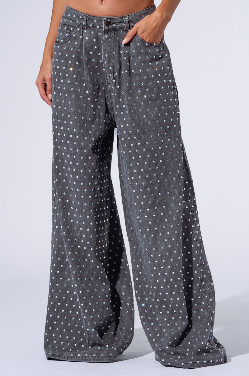 A person is modeling high-waisted, wide-leg black denim pants named "Dazzled Rhinestone Studded Denim Pants," featuring subtle silver rhinestone embellishments. Their hand is casually placed in their pocket, and the background is plain and light-colored.