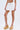 A woman wears the LUCKY YOU EYELET MINI SKIRT, a short white high-rise mini skirt, paired with beige ankle-strap platform wedges. The image is cropped from shoulders down against a plain light background.