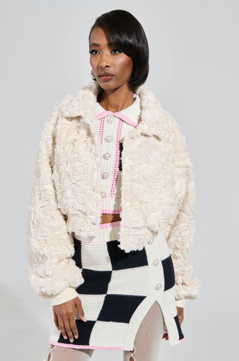 A person stands wearing the FLORA TEXTURED BOMBER, an elegant faux fur jacket in cream, paired with a checkered skirt featuring black-and-white tones and pink accents. The cropped silhouette contrasts against the plain gray background, emphasizing the ensemble's modern elegance.